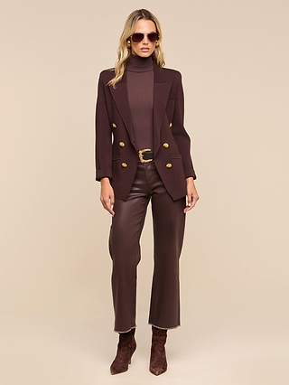 Woman wearing brown sunglasses, a dark brown turtleneck, the Amelia Knit Blazer with gold buttons and removable shoulder pads, brown pants with a belt, and brown ankle boots, standing against a beige background.