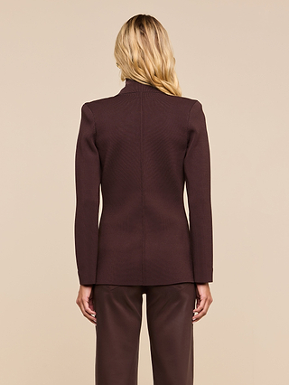 A person with long blonde hair stands facing away, showing the back of their outfit—a dark brown Amelia Knit Blazer with removable shoulder pads and matching pants—against a plain beige background.