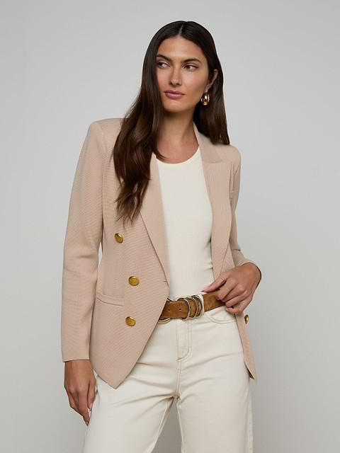A woman with long brown hair wears the Amelia Knit Blazer, featuring gold buttons, styled over a white top and cream pants. She stands against a plain background with one hand in her pocket, gazing to the side.