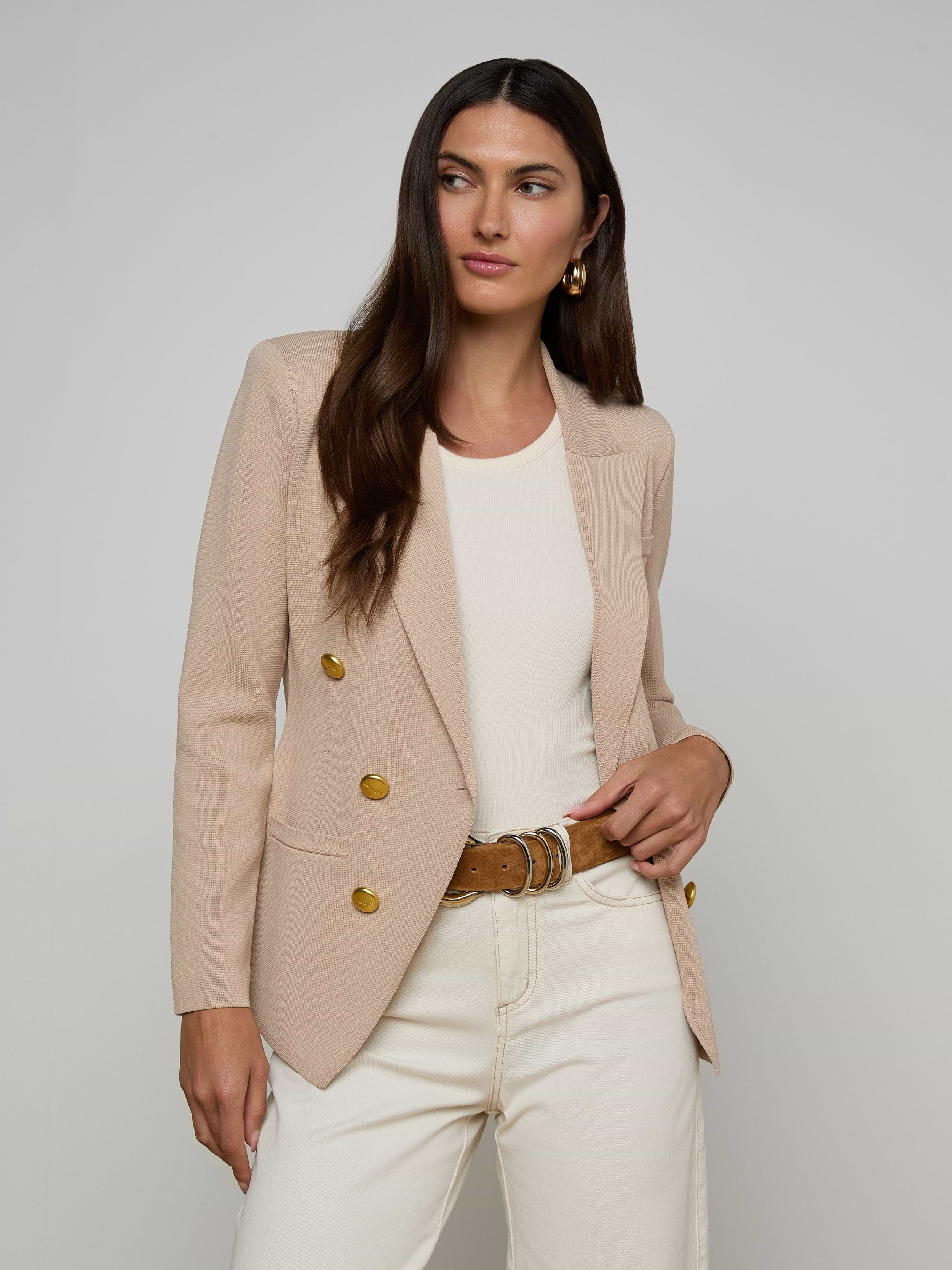 A woman with long brown hair wears the Amelia Knit Blazer, featuring gold buttons, styled over a white top and cream pants. She stands against a plain background with one hand in her pocket, gazing to the side.
