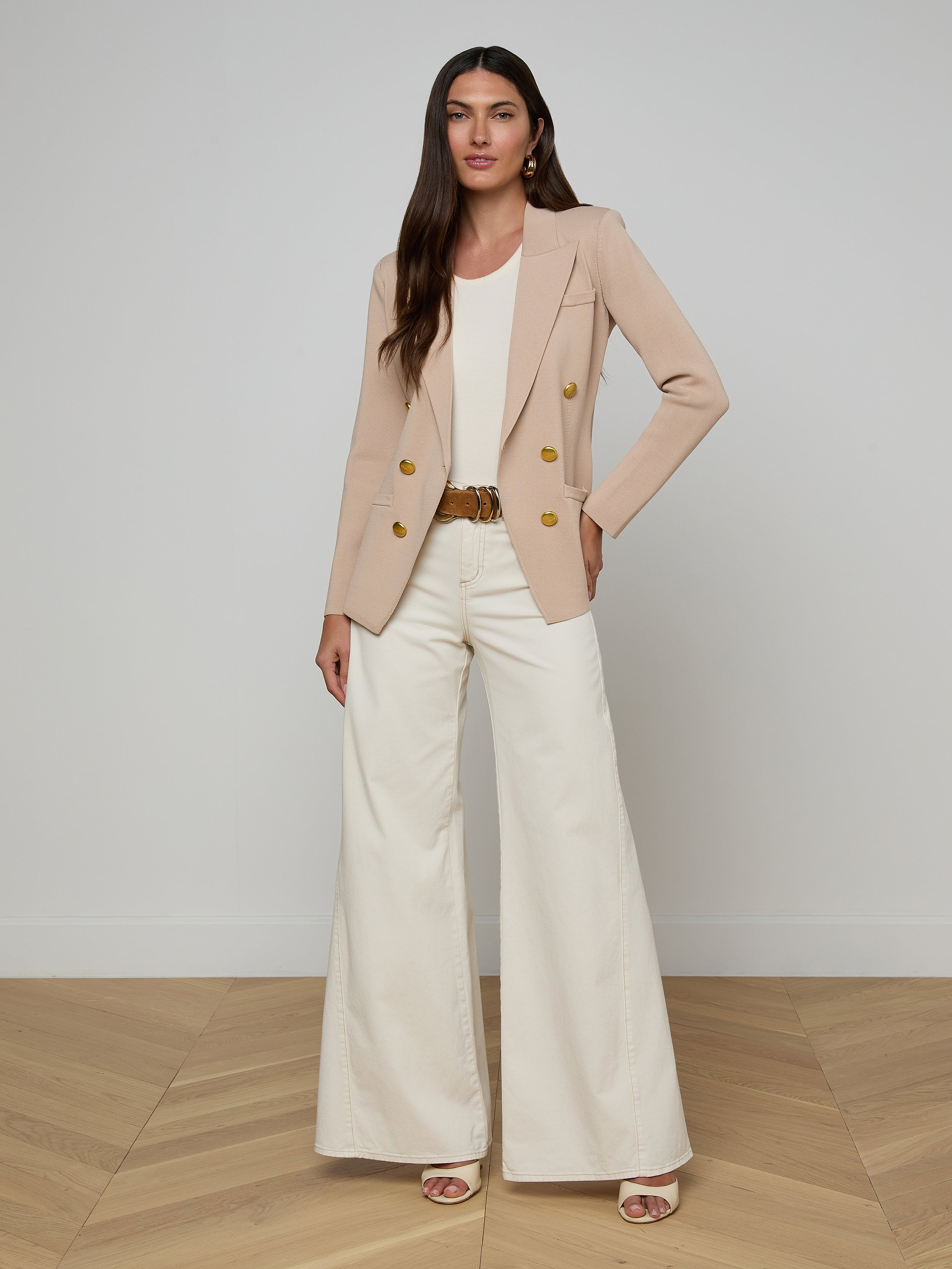 A woman stands against a plain white background, posing with one hand on her hip and the other relaxed by her side, dressed in the Amelia Knit Blazer with gold buttons, a white top, and wide-leg white pants.