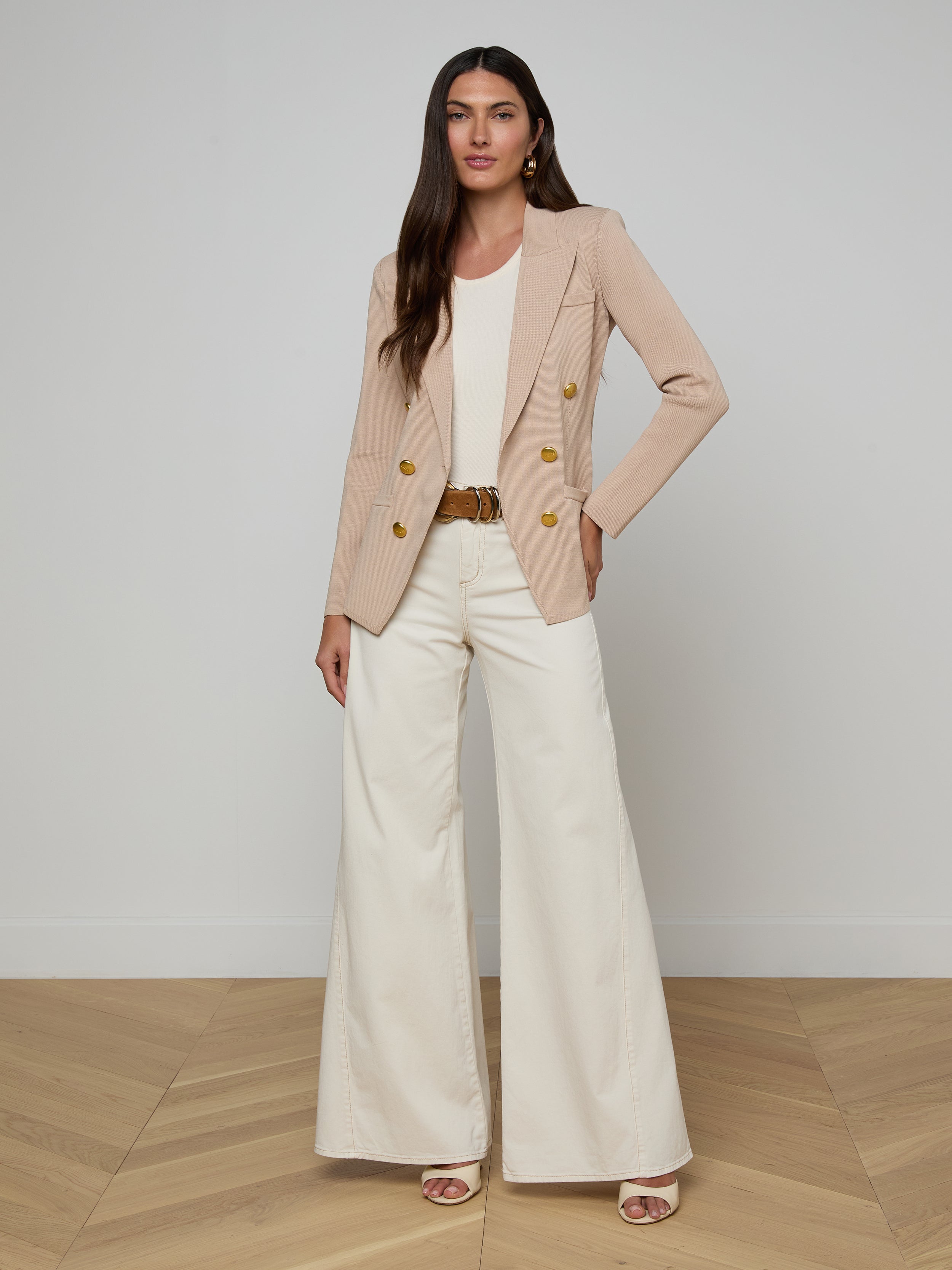 A woman stands against a plain white background, posing with one hand on her hip and the other relaxed by her side, dressed in the Amelia Knit Blazer with gold buttons, a white top, and wide-leg white pants.