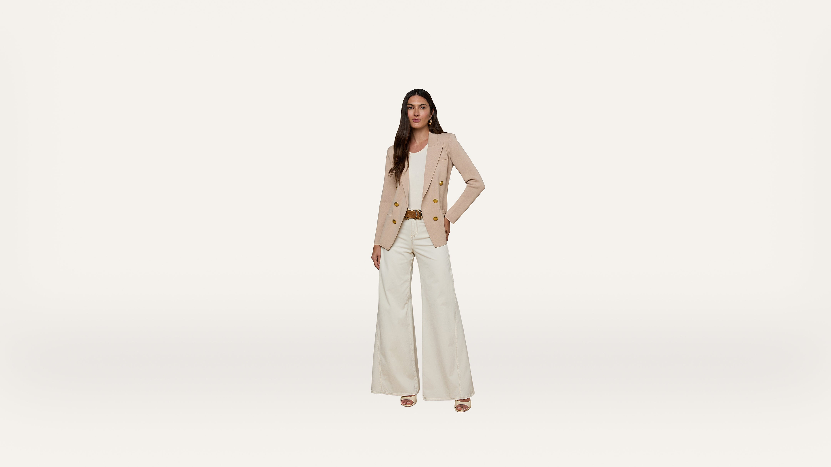Woman standing against a plain white background, wearing a beige blazer, light pink blouse, white wide-leg pants, and beige heels, with one hand on her hip and a neutral expression.