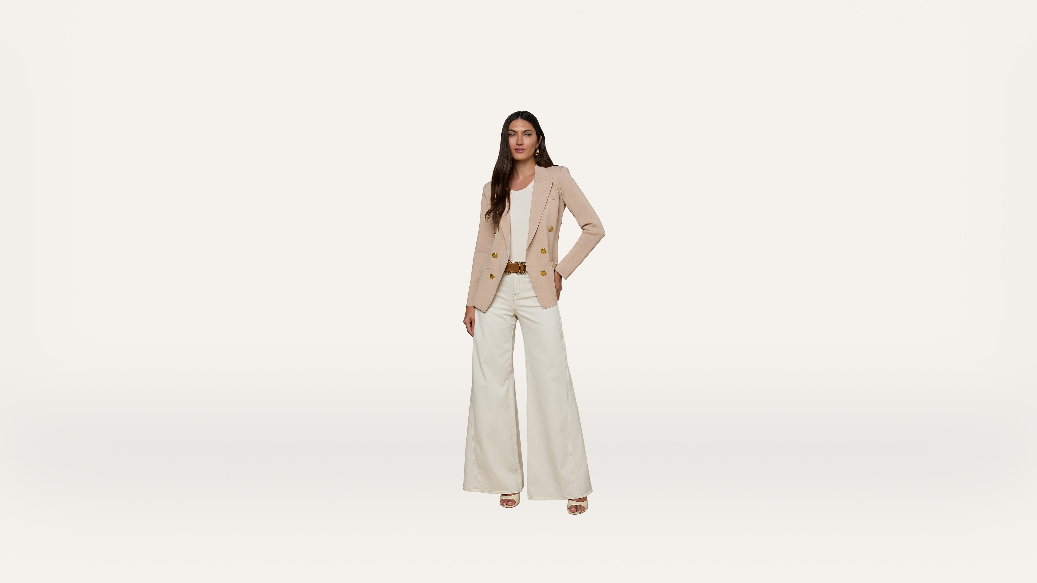 Woman standing against a plain white background, wearing a beige blazer, light pink blouse, white wide-leg pants, and beige heels, with one hand on her hip and a neutral expression.
