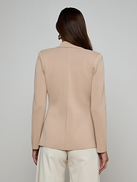 A woman with long brown hair stands facing away from the camera against a plain light background, wearing the Amelia Knit Blazer—a beige structured knit blazer with gold buttons—paired with white pants.