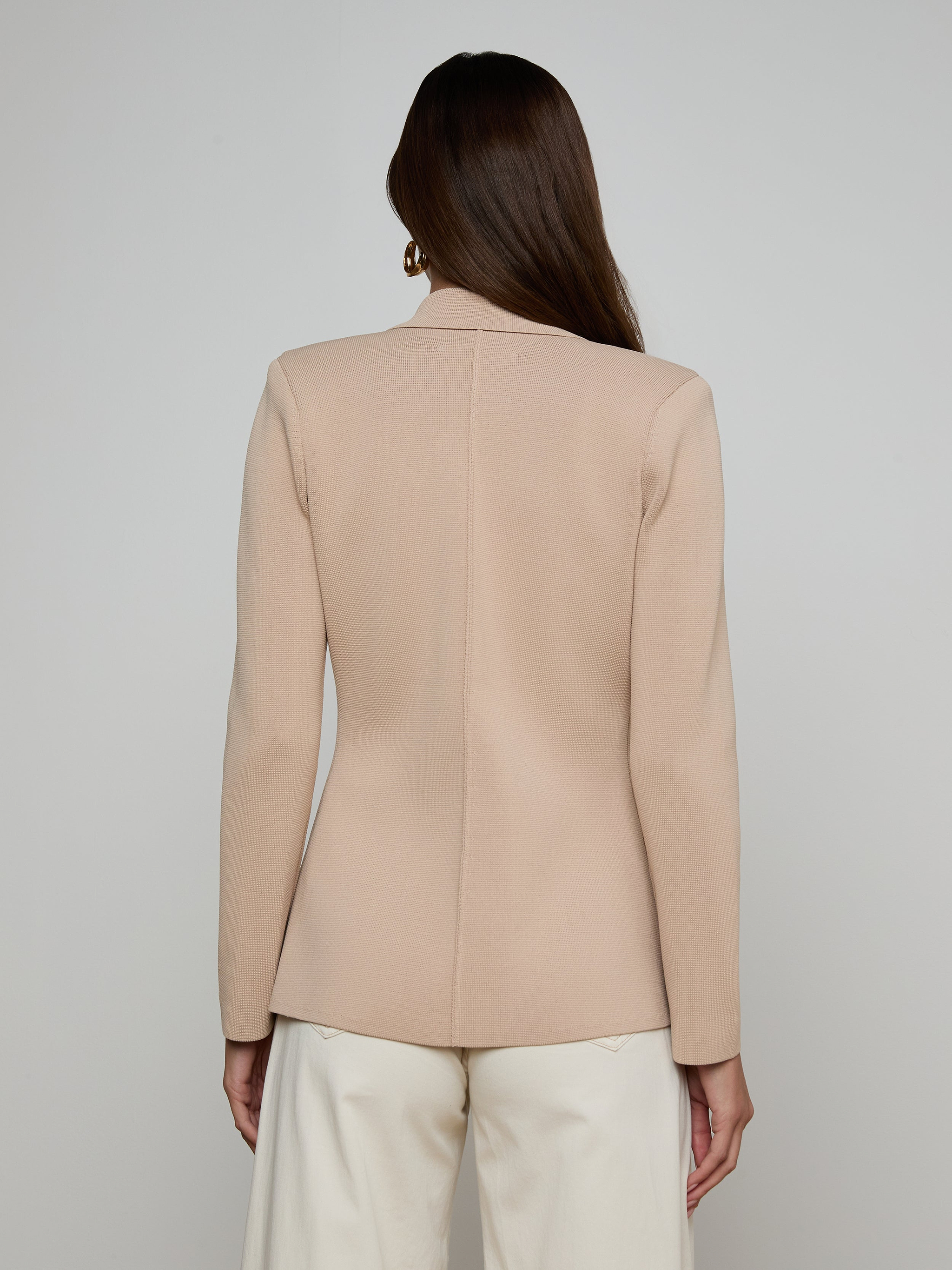 A woman with long brown hair stands facing away from the camera against a plain light background, wearing the Amelia Knit Blazer—a beige structured knit blazer with gold buttons—paired with white pants.