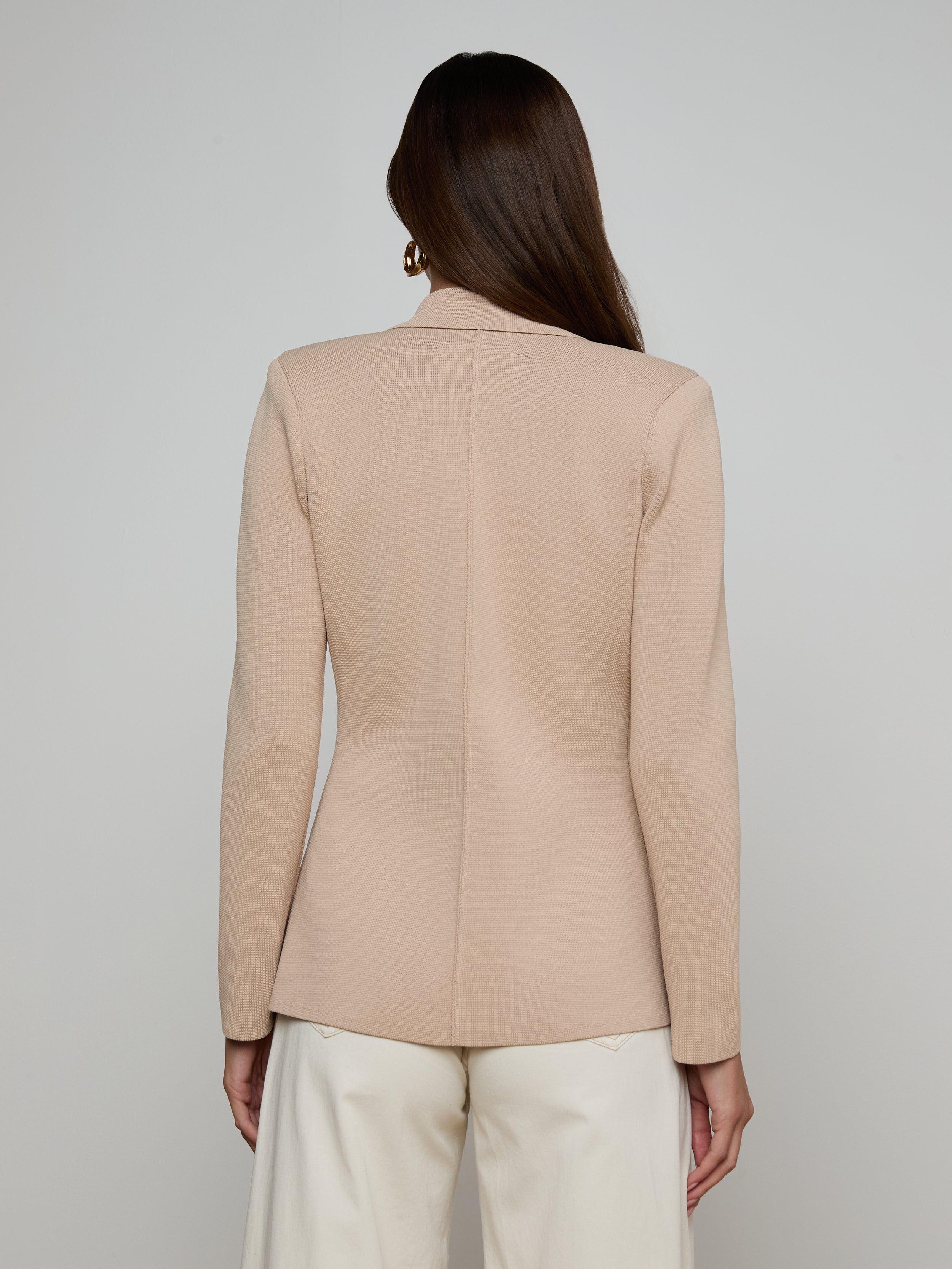 A woman with long brown hair stands facing away from the camera against a plain light background, wearing the Amelia Knit Blazer—a beige structured knit blazer with gold buttons—paired with white pants.