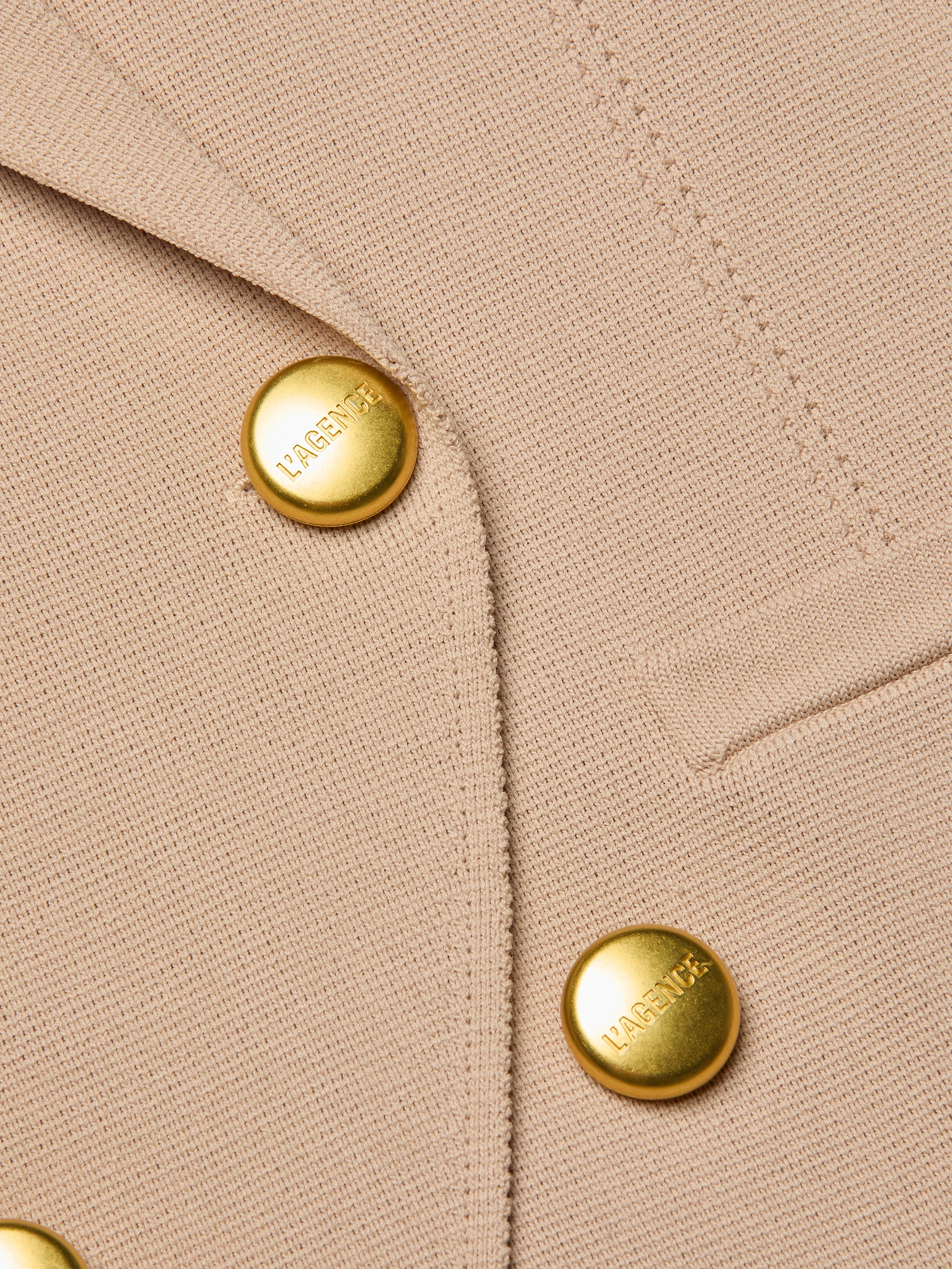 Close-up of the Amelia Knit Blazer, crafted from beige textured knit and detailed with two shiny gold L’AGENCE-engraved buttons and a precisely stitched small welt pocket.