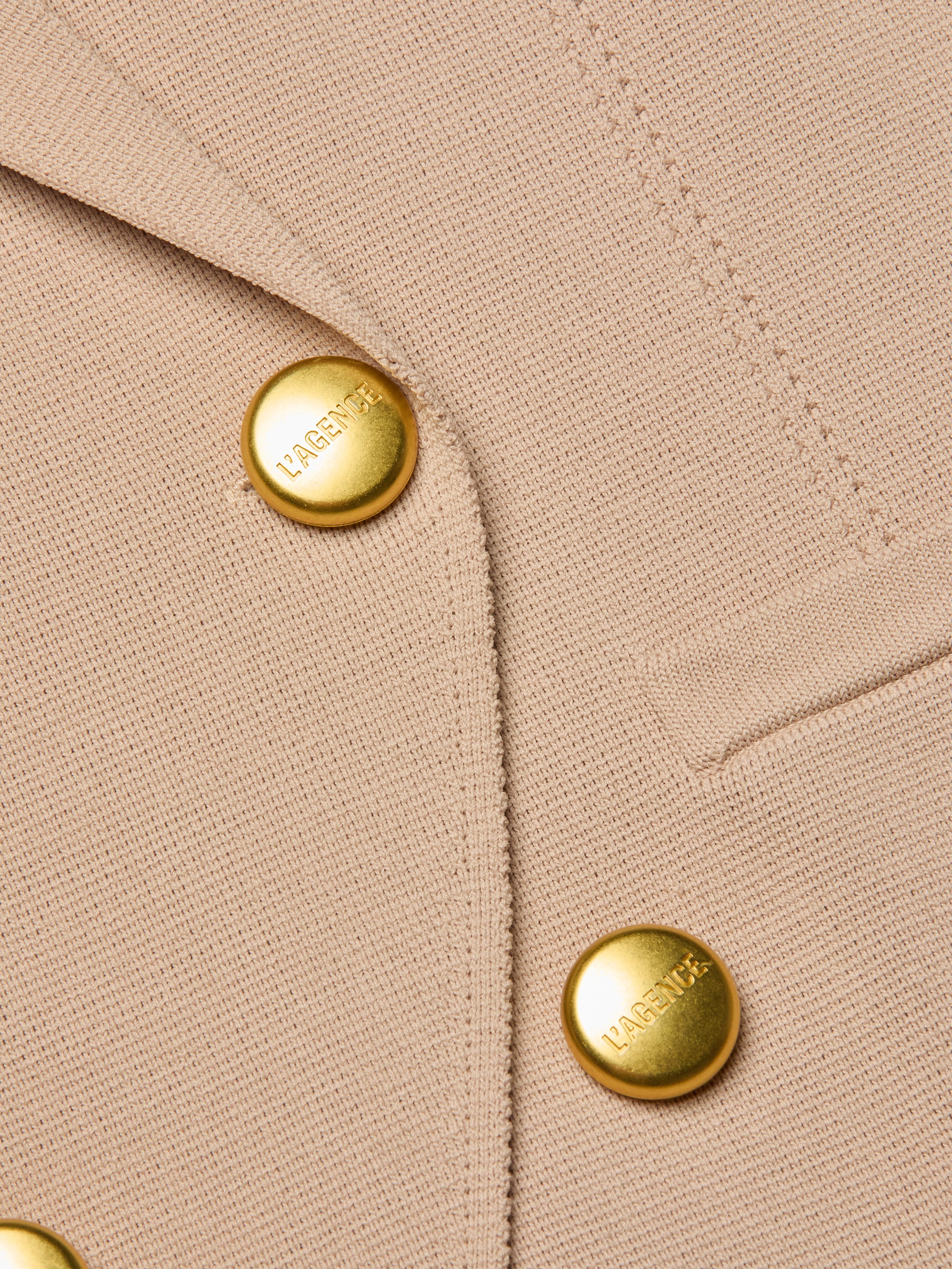 Close-up of the Amelia Knit Blazer, crafted from beige textured knit and detailed with two shiny gold L’AGENCE-engraved buttons and a precisely stitched small welt pocket.