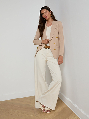 A woman with long brown hair and a neutral expression stands against a white wall wearing the Amelia Knit Blazer in beige with gold buttons, paired with a white top, cream wide-leg pants, a brown belt, and beige heels.