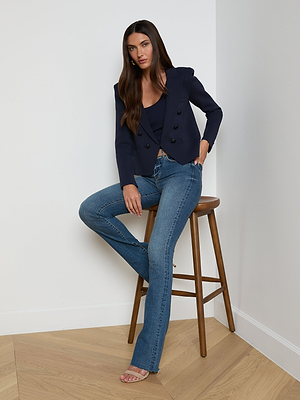 A woman with long dark hair, dressed in the Mercer Knit Blazer, dark top, blue jeans, and heeled sandals, sits on a wooden stool in a minimalist room with white walls and light wood flooring.