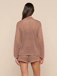 A woman is shown from behind wearing the Mariana Pointelle Knit Shirt, a light brown airy long-sleeve top paired with matching see-through shorts. The background is plain off-white.