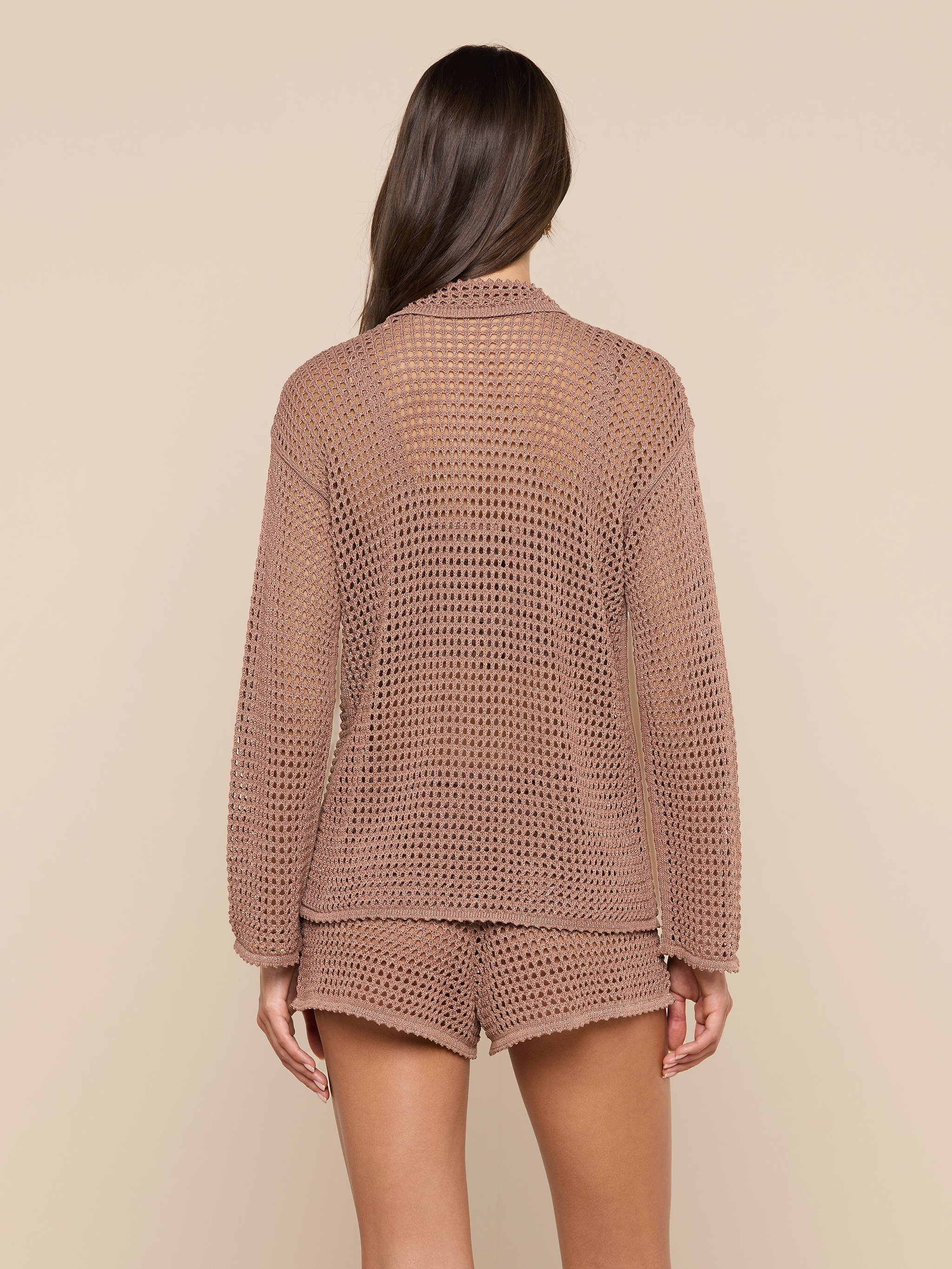 A woman is shown from behind wearing the Mariana Pointelle Knit Shirt, a light brown airy long-sleeve top paired with matching see-through shorts. The background is plain off-white.