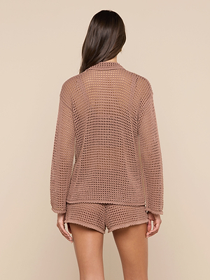A woman faces away, wearing the Mariana Pointelle Knit Shirt in light brown with a net-like mesh texture and matching shorts—an ideal vacation set. The background is solid beige.