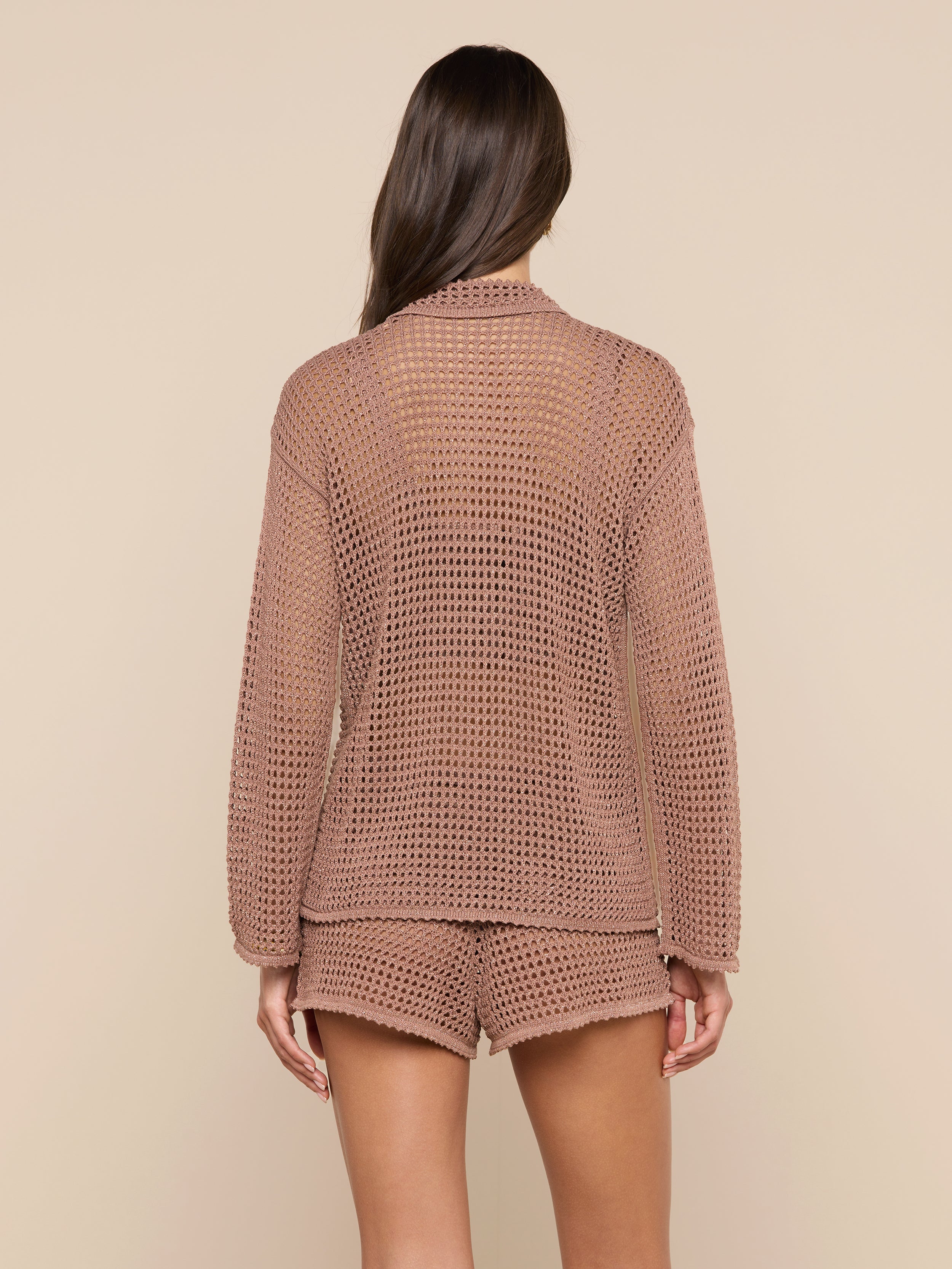 A woman is shown from behind wearing the Mariana Pointelle Knit Shirt, a light brown airy long-sleeve top paired with matching see-through shorts. The background is plain off-white.