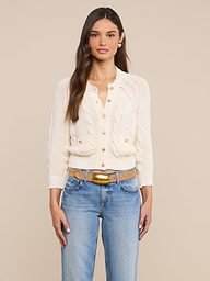 A woman with long brown hair wears the Darcy Cable Knit Cardigan—a cream-colored cotton-blend knit button-up cardigan with ruffle details—paired with blue jeans, standing against a plain light background.