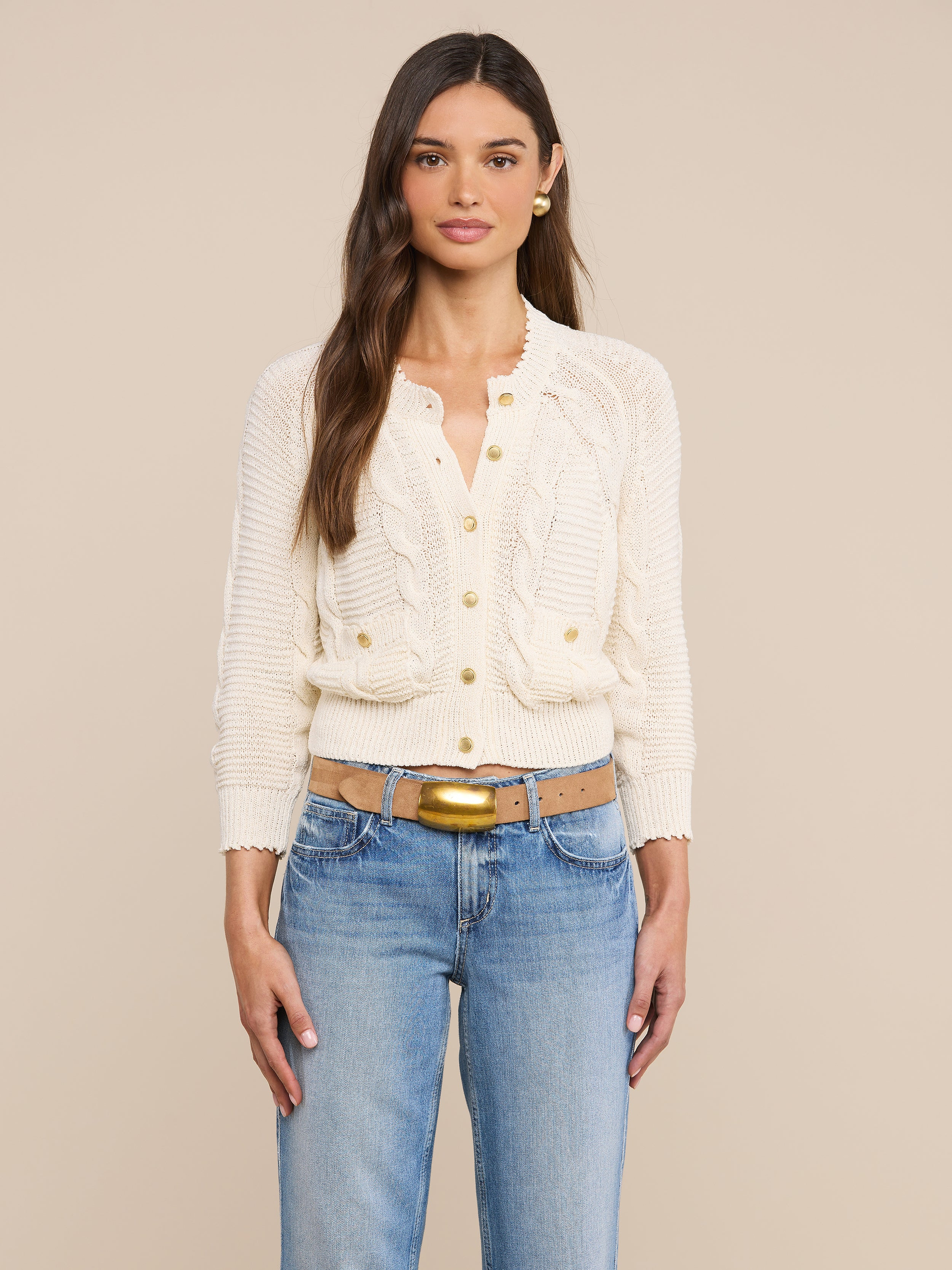 A woman with long brown hair wears the Darcy Cable Knit Cardigan—a cream-colored cotton-blend knit button-up cardigan with ruffle details—paired with blue jeans, standing against a plain light background.