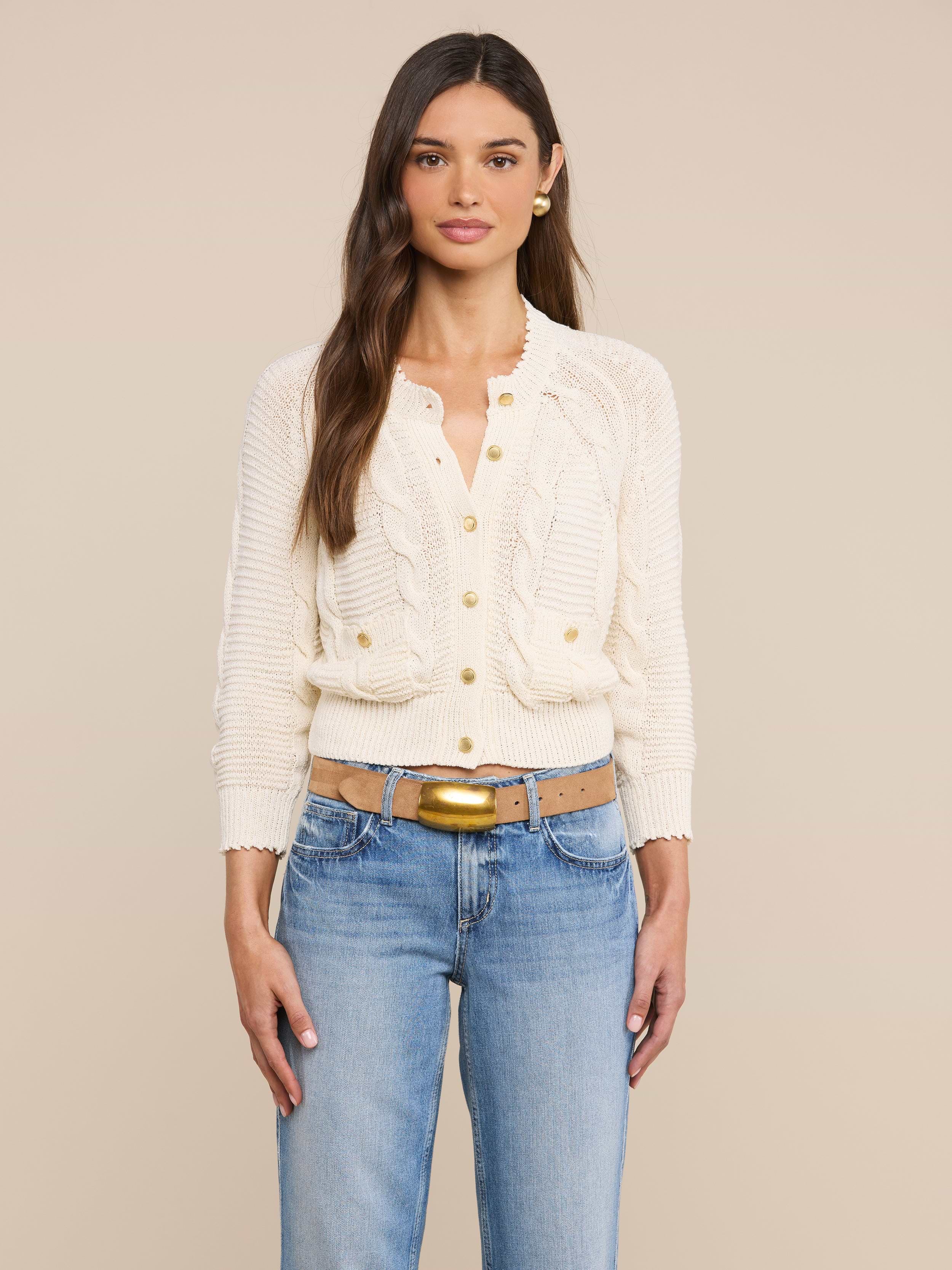 A woman with long brown hair wears the Darcy Cable Knit Cardigan—a cream-colored cotton-blend knit button-up cardigan with ruffle details—paired with blue jeans, standing against a plain light background.