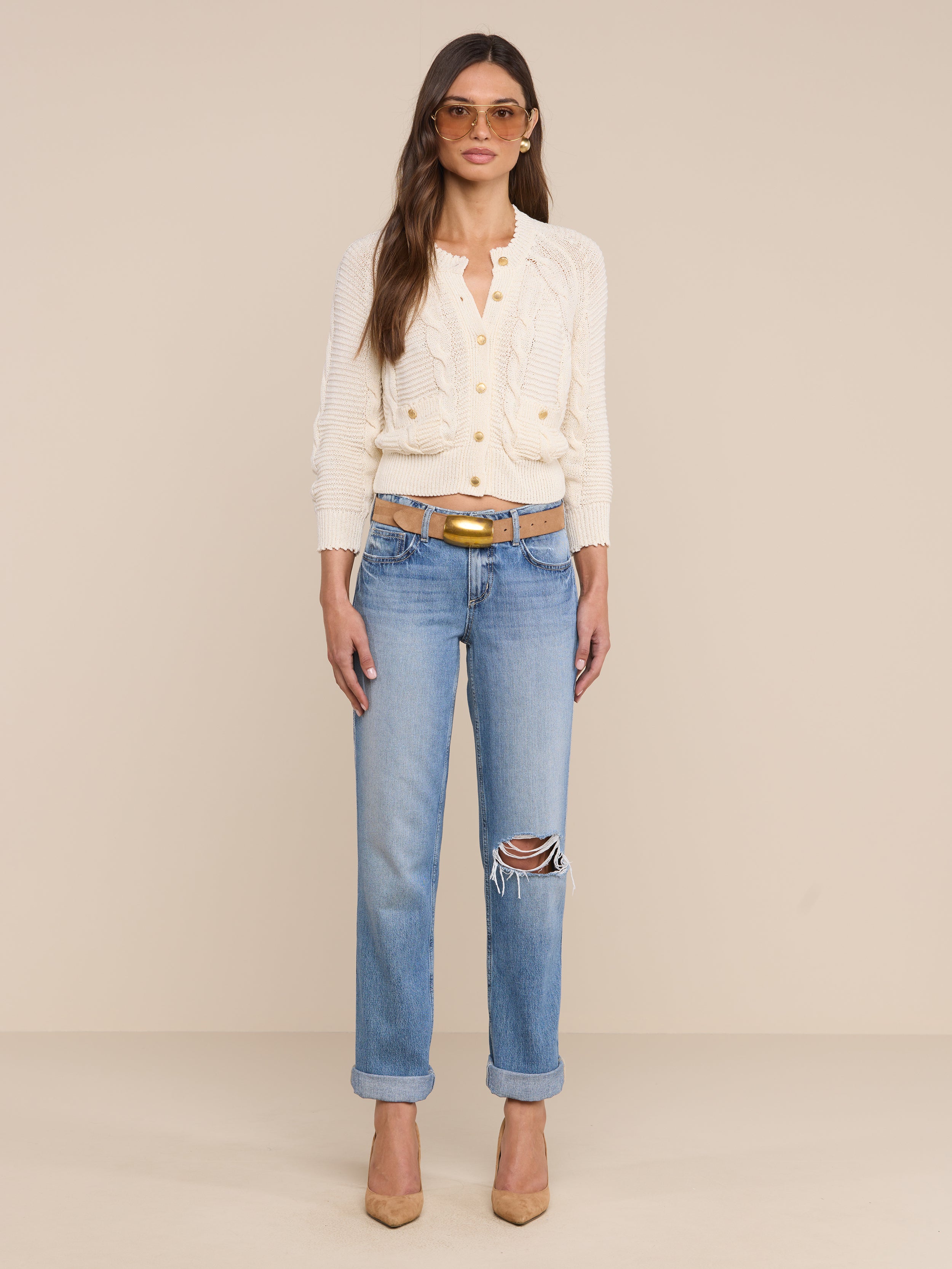 A woman wearing sunglasses, the Darcy Cable Knit Cardigan in white with patch pockets, blue ripped jeans with a brown belt, and beige heels stands against a plain white background.