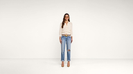 A woman wearing sunglasses, a white blouse, blue ripped jeans, a brown belt, and tan heels stands in a white, minimalist room, facing forward.