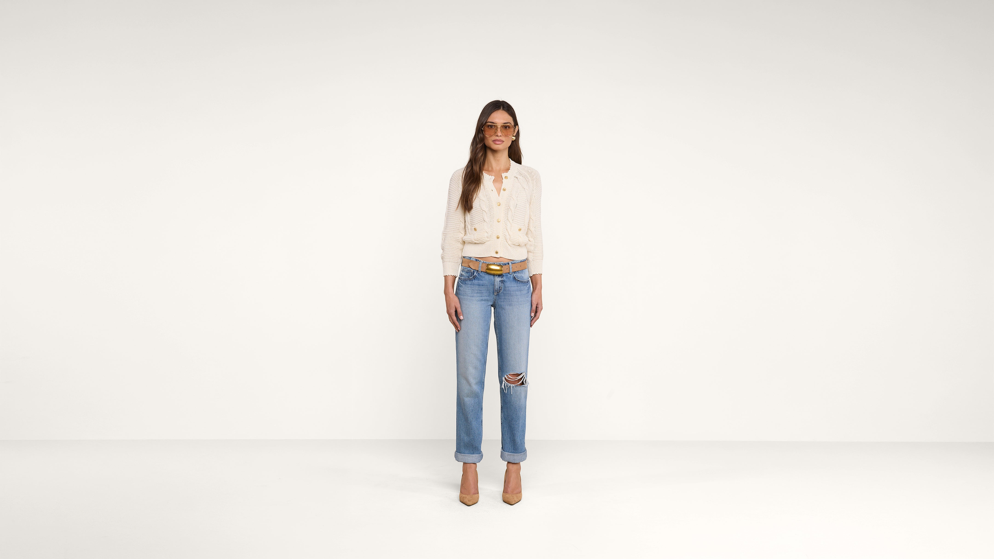 A woman wearing sunglasses, a white blouse, blue ripped jeans, a brown belt, and tan heels stands in a white, minimalist room, facing forward.