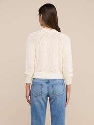 A person with long brown hair, wearing the Darcy Cable Knit Cardigan in a cream-colored cotton-blend, paired with blue jeans, is shown from the back against a plain light background.