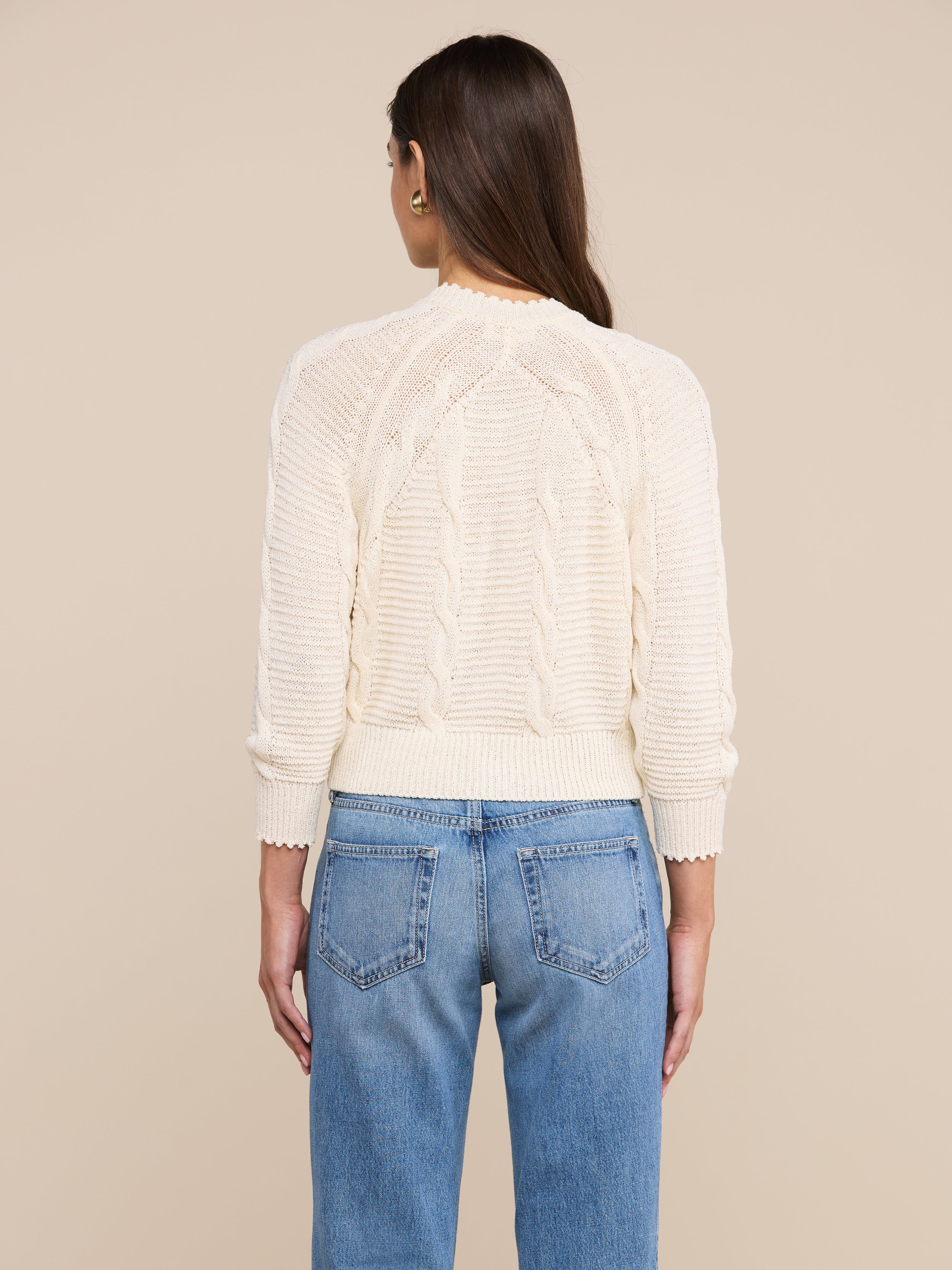 A person with long brown hair, wearing the Darcy Cable Knit Cardigan in a cream-colored cotton-blend, paired with blue jeans, is shown from the back against a plain light background.