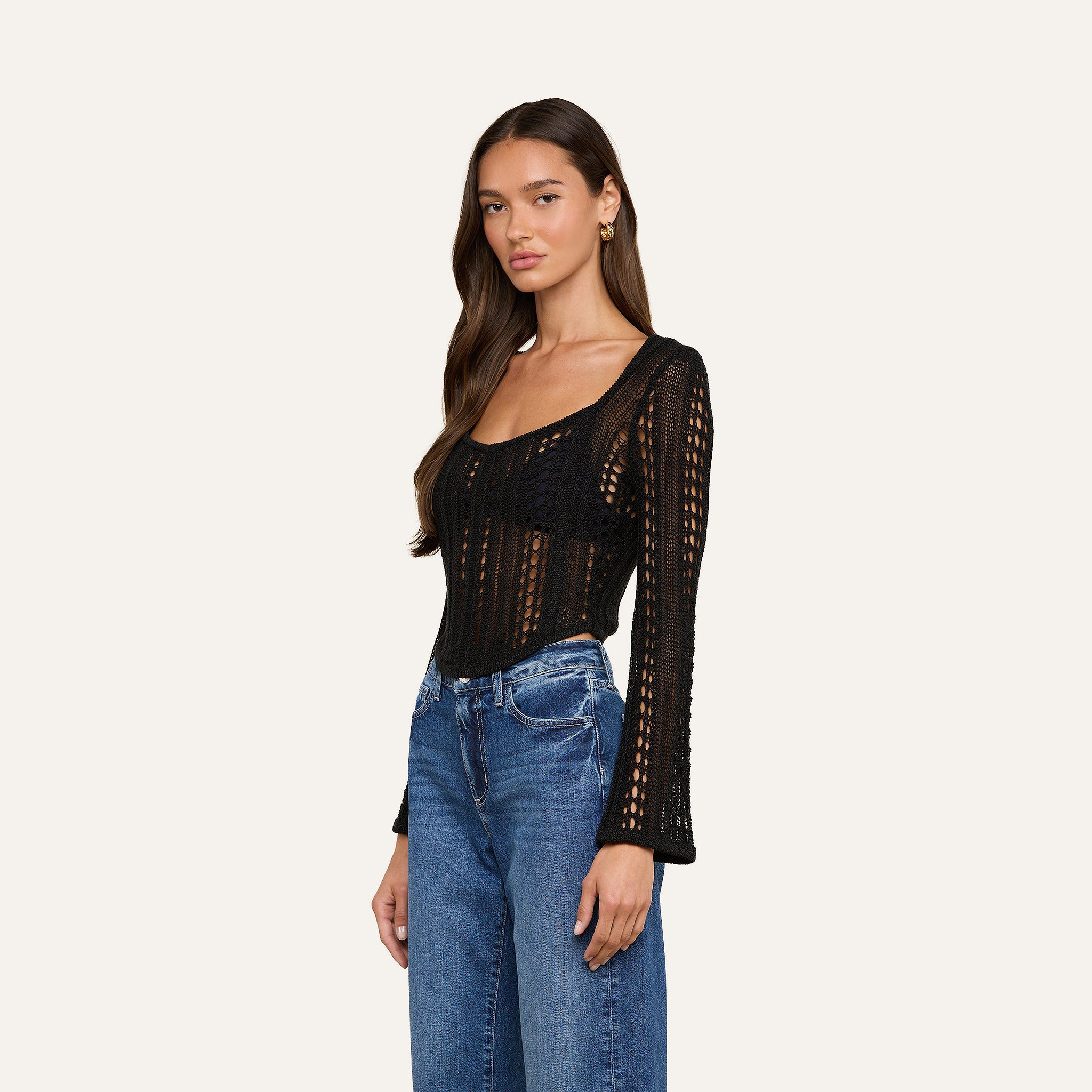 A woman wearing the Orine Pointelle Knit Top in black, featuring long sleeves and a scoop neckline, paired with blue jeans and standing against a plain off-white background.