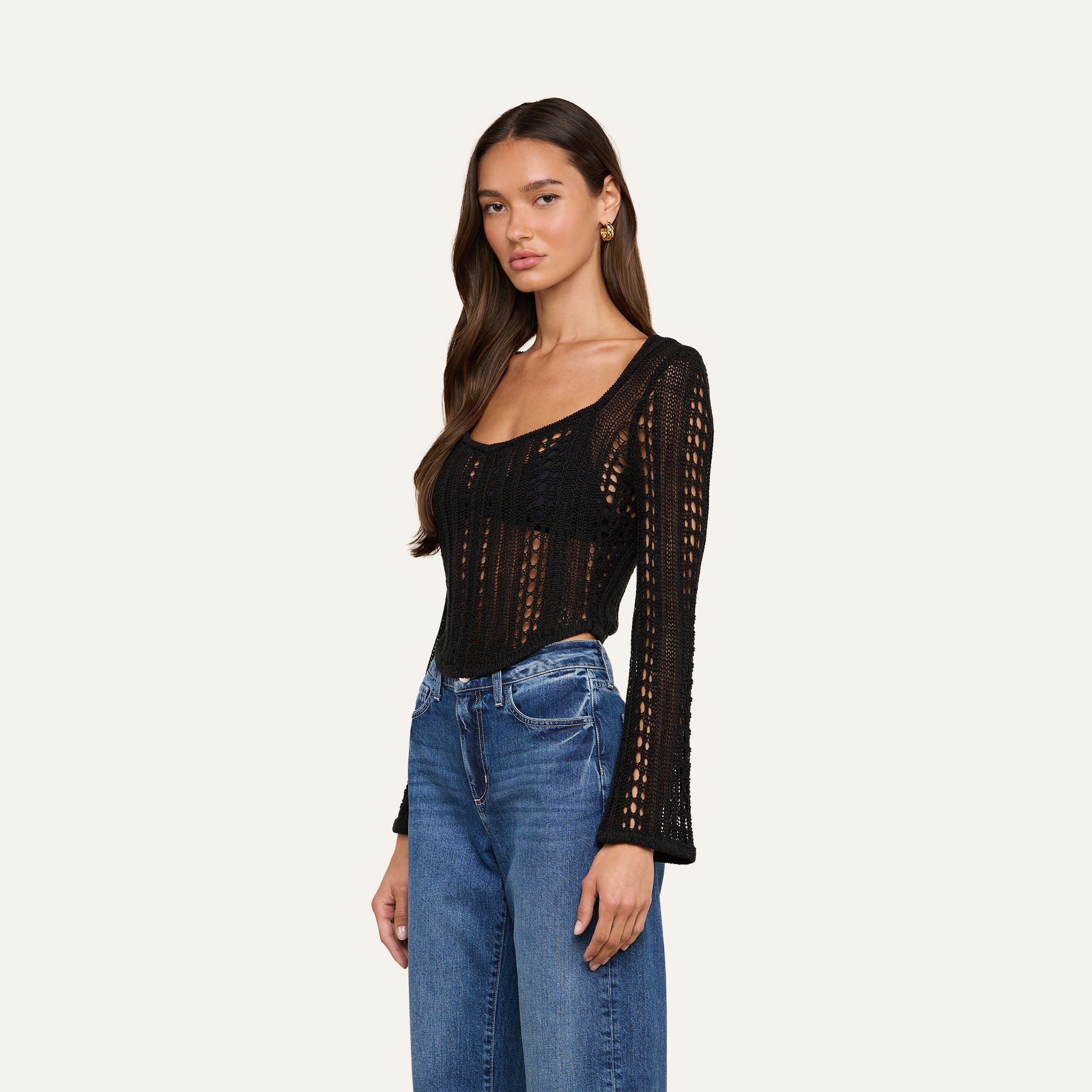 A woman wearing the Orine Pointelle Knit Top in black, featuring long sleeves and a scoop neckline, paired with blue jeans and standing against a plain off-white background.