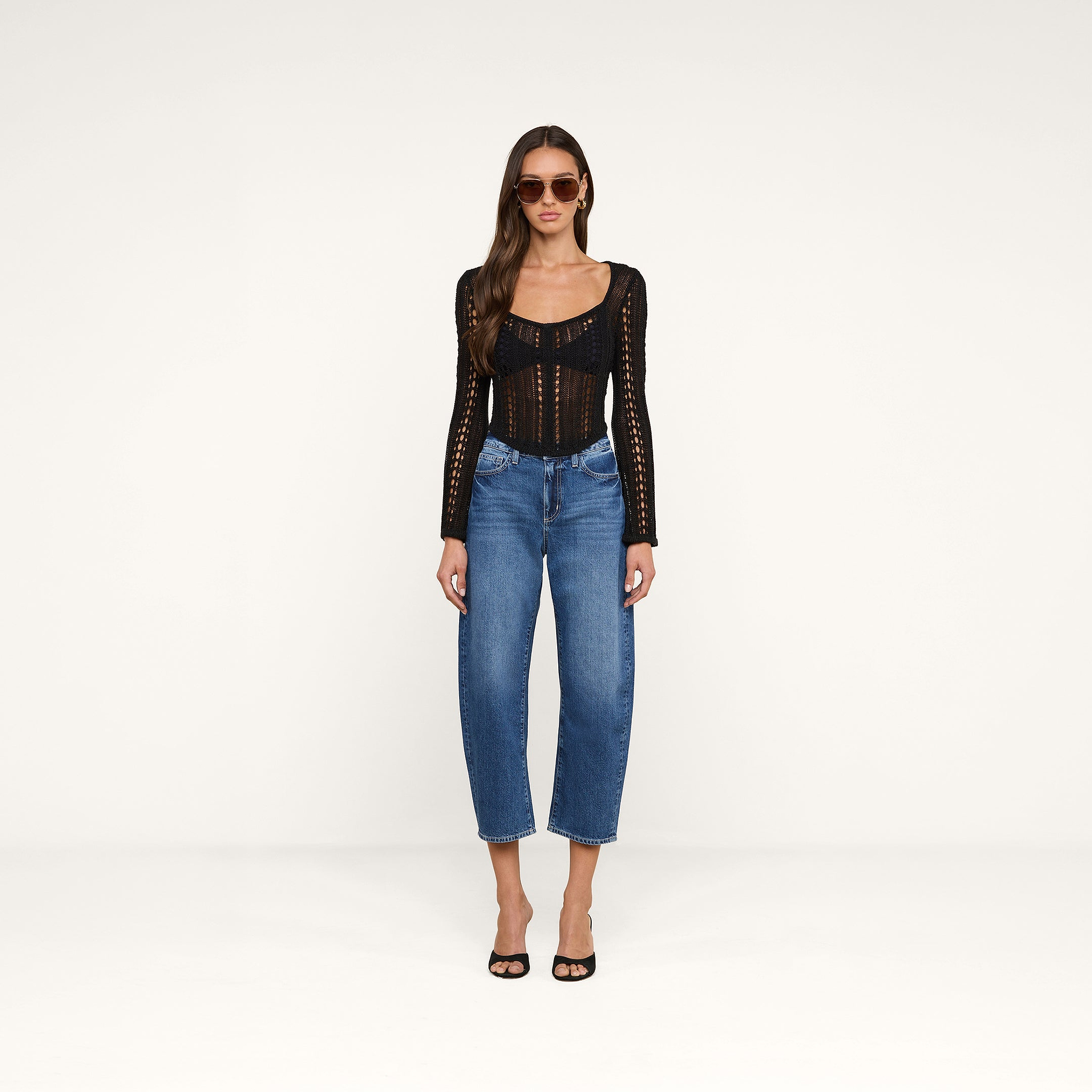 A woman wearing sunglasses, the Orine Pointelle Knit Top with bell sleeves, blue high-waisted jeans, and black open-toe heels stands against a plain white background.