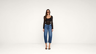 Woman wearing sunglasses, a black long-sleeved top, blue jeans, and black heels stands against a plain white background.