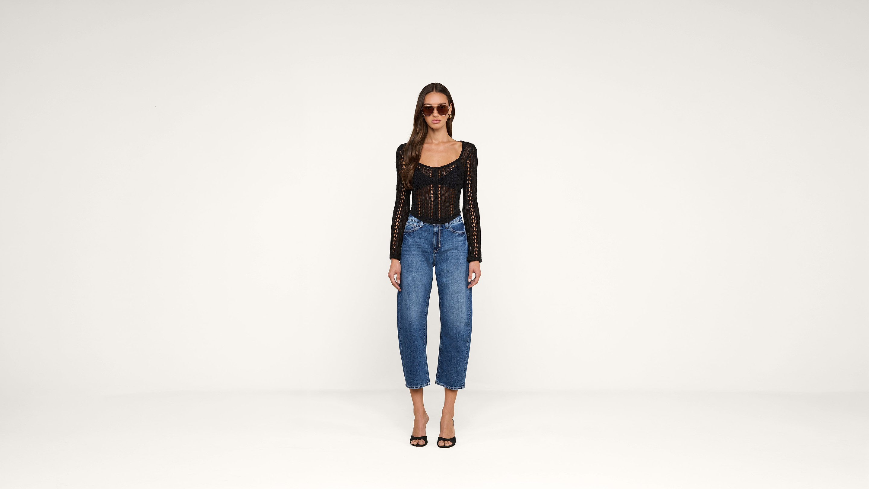 Woman wearing sunglasses, a black long-sleeved top, blue jeans, and black heels stands against a plain white background.