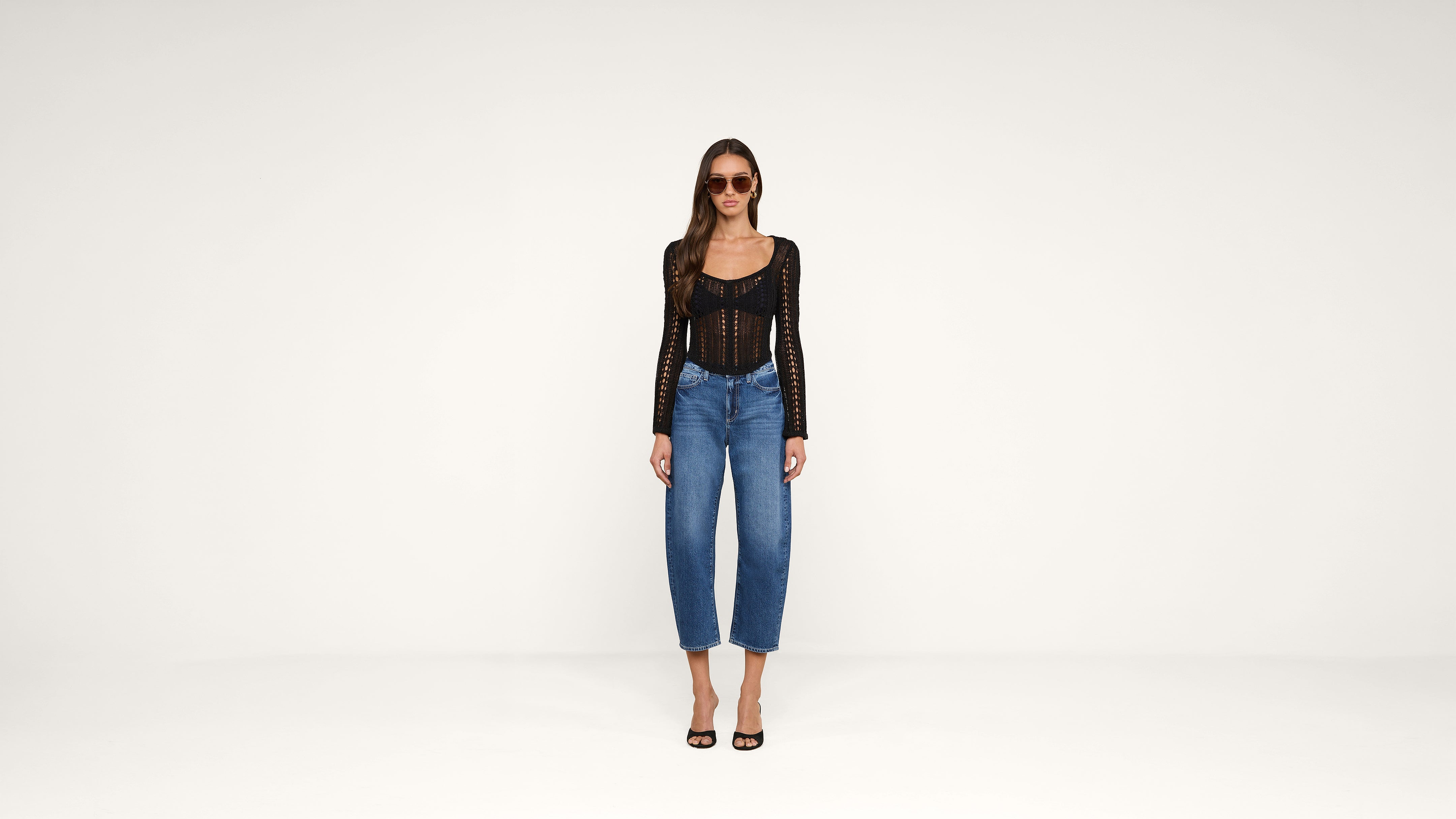 Woman wearing sunglasses, a black long-sleeved top, blue jeans, and black heels stands against a plain white background.