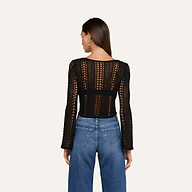 A woman with long brown hair, seen from behind, is wearing the Orine Pointelle Knit Top — a black, long-sleeve top featuring an open knit pattern — paired with blue high-waisted jeans. She stands against a plain, light background.