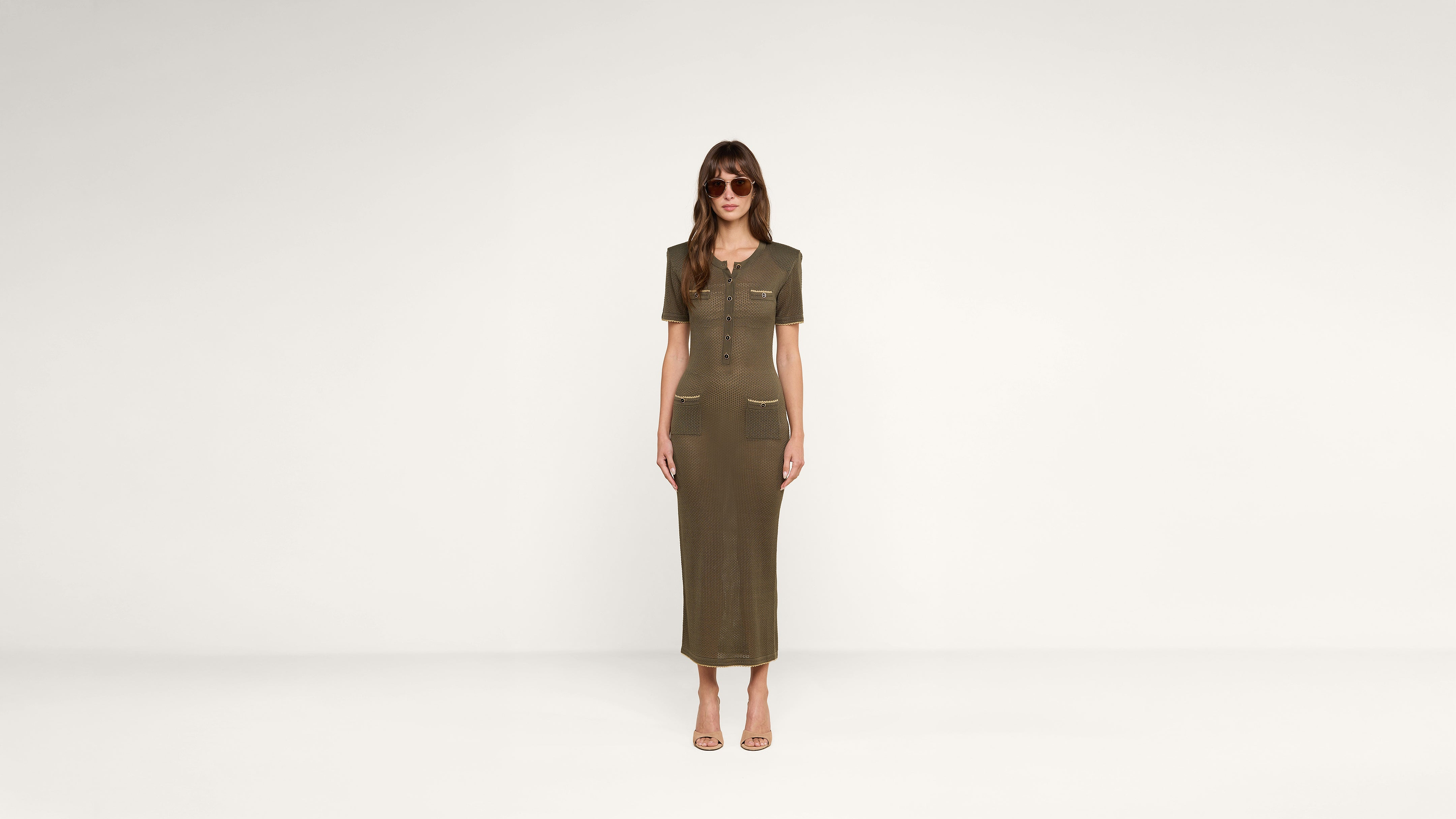 Woman standing against a plain white background, wearing sunglasses and a short-sleeved, olive green, button-front, midi-length dress with front pockets, and beige open-toe heels.