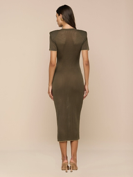 A woman stands facing away, wearing the Mel Knit Dress—a fitted midi dress with shoulder pads in olive green—paired with beige high heels against a plain white background.