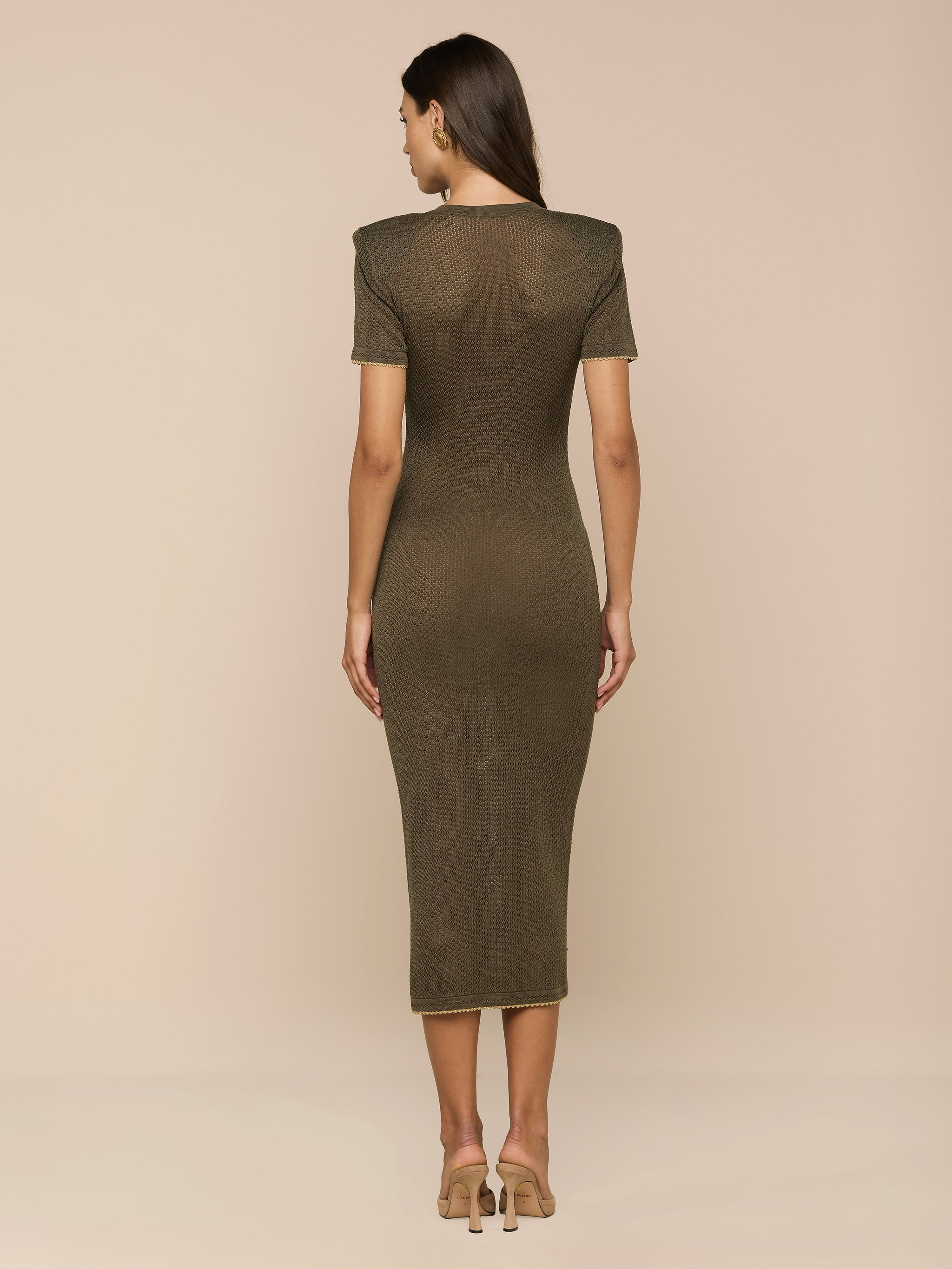 A woman stands facing away, wearing the Mel Knit Dress—a fitted midi dress with shoulder pads in olive green—paired with beige high heels against a plain white background.