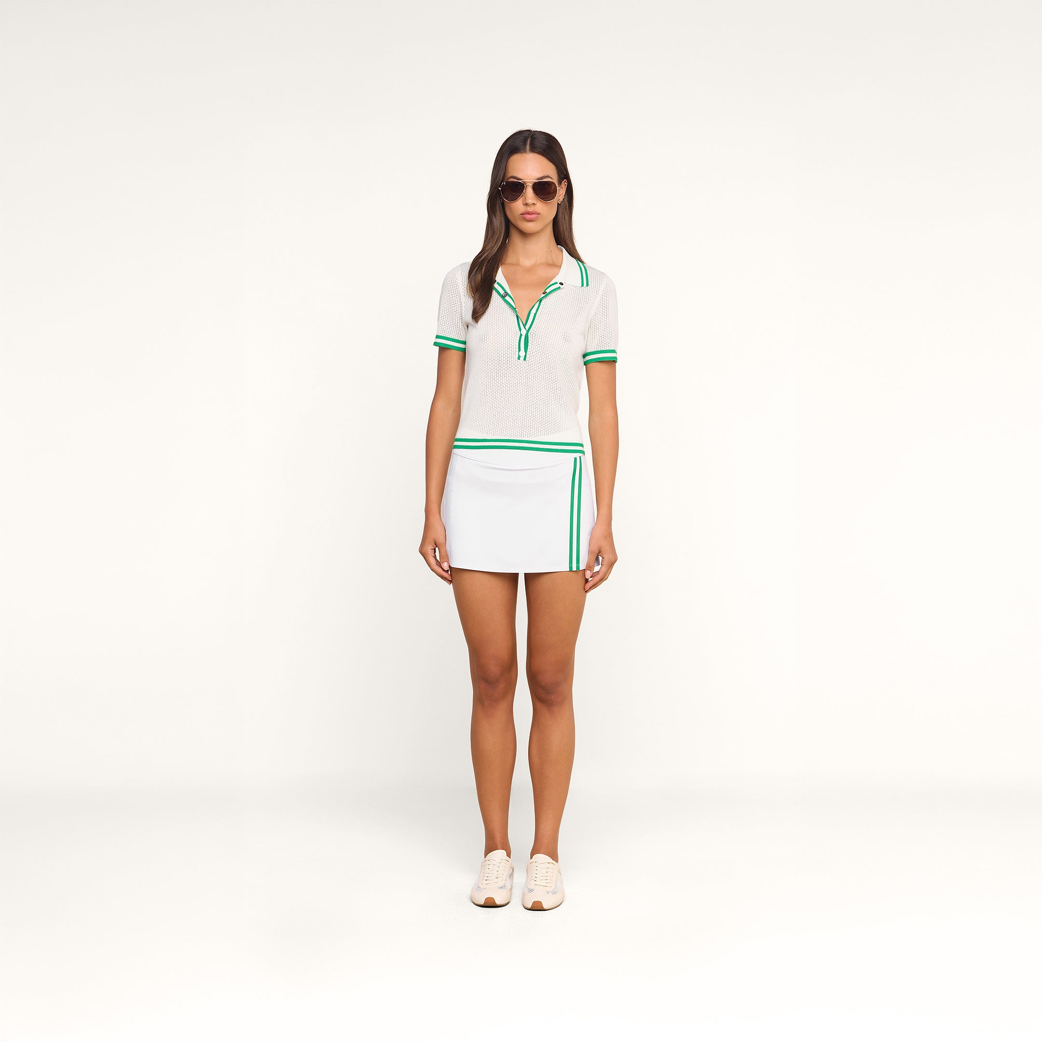A woman wearing sunglasses, the Calvin Polo Top—a featherweight knit tennis polo with green trim—and a white skirt with green stripes stands facing forward against a plain white background.
