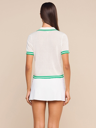 A woman stands facing away, wearing the Calvin Polo Top—a featherweight white knit mesh top with green trim—and a white tennis skirt against a beige background.