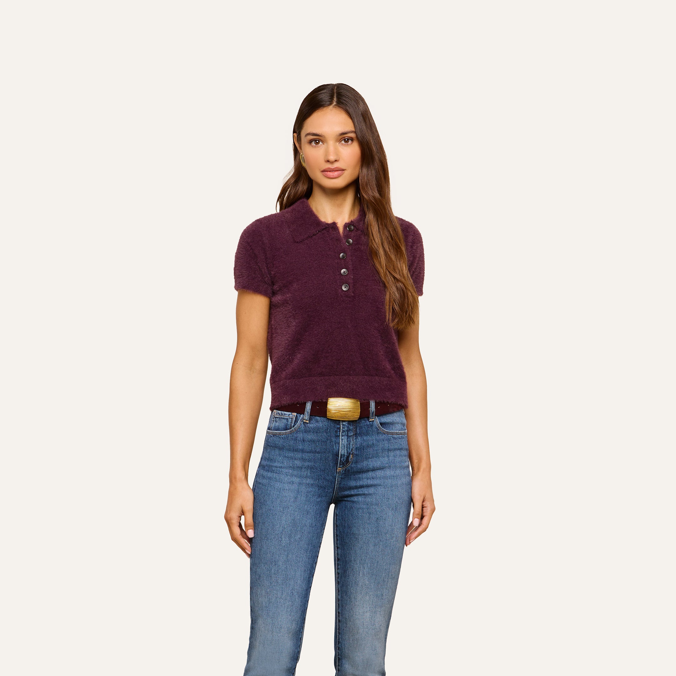 A woman with long brown hair wears the Cirrus Cropped Polo—a short-sleeved, plush dark purple knit collared shirt—paired with blue jeans. She stands facing forward against a plain, light background, her expression neutral and arms relaxed at her sides.