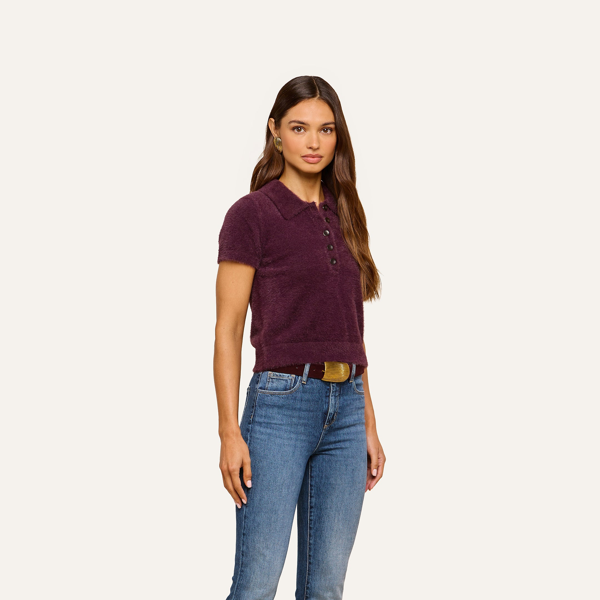 A woman with long brown hair wears the Cirrus Cropped Polo, crafted from plush dark purple knit, paired with blue jeans. She stands facing forward with a neutral expression against a plain, light background.
