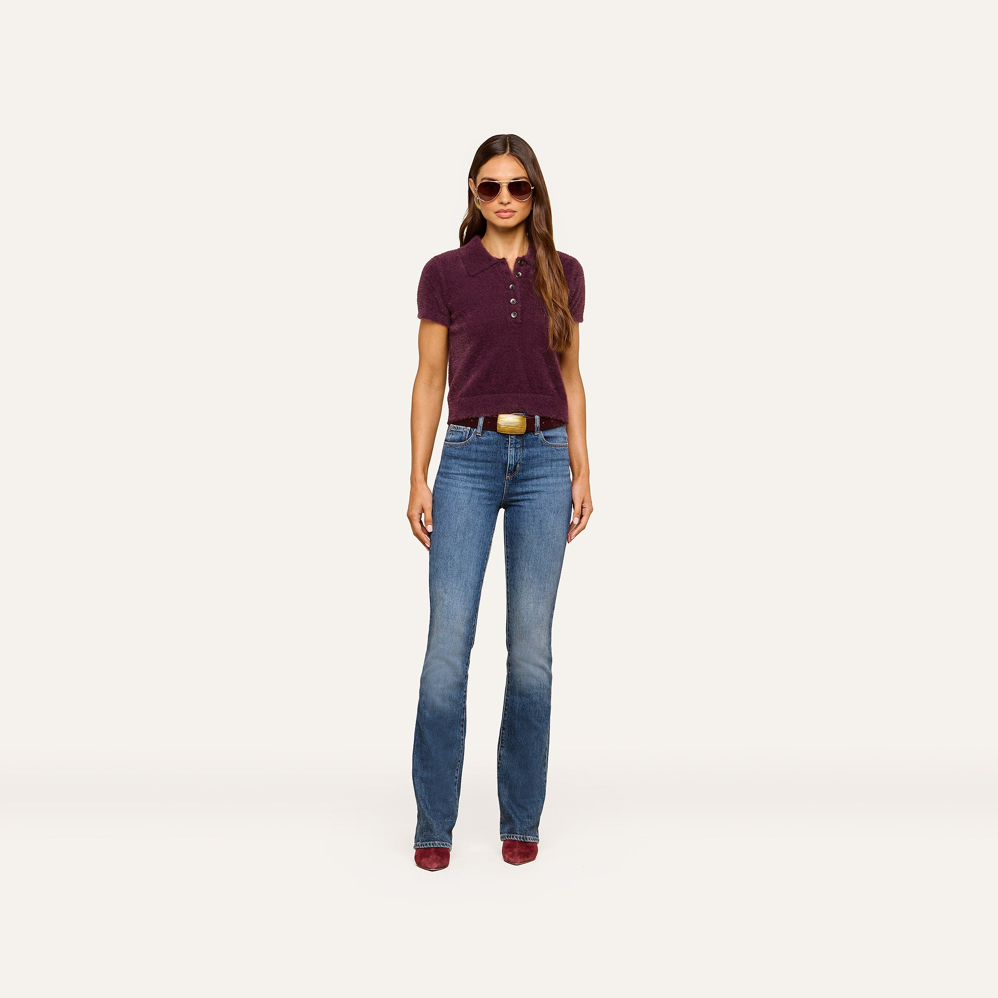 A woman stands facing forward against a plain white background, wearing sunglasses, blue jeans with a belt, red shoes, and the Cirrus Cropped Polo in maroon.
