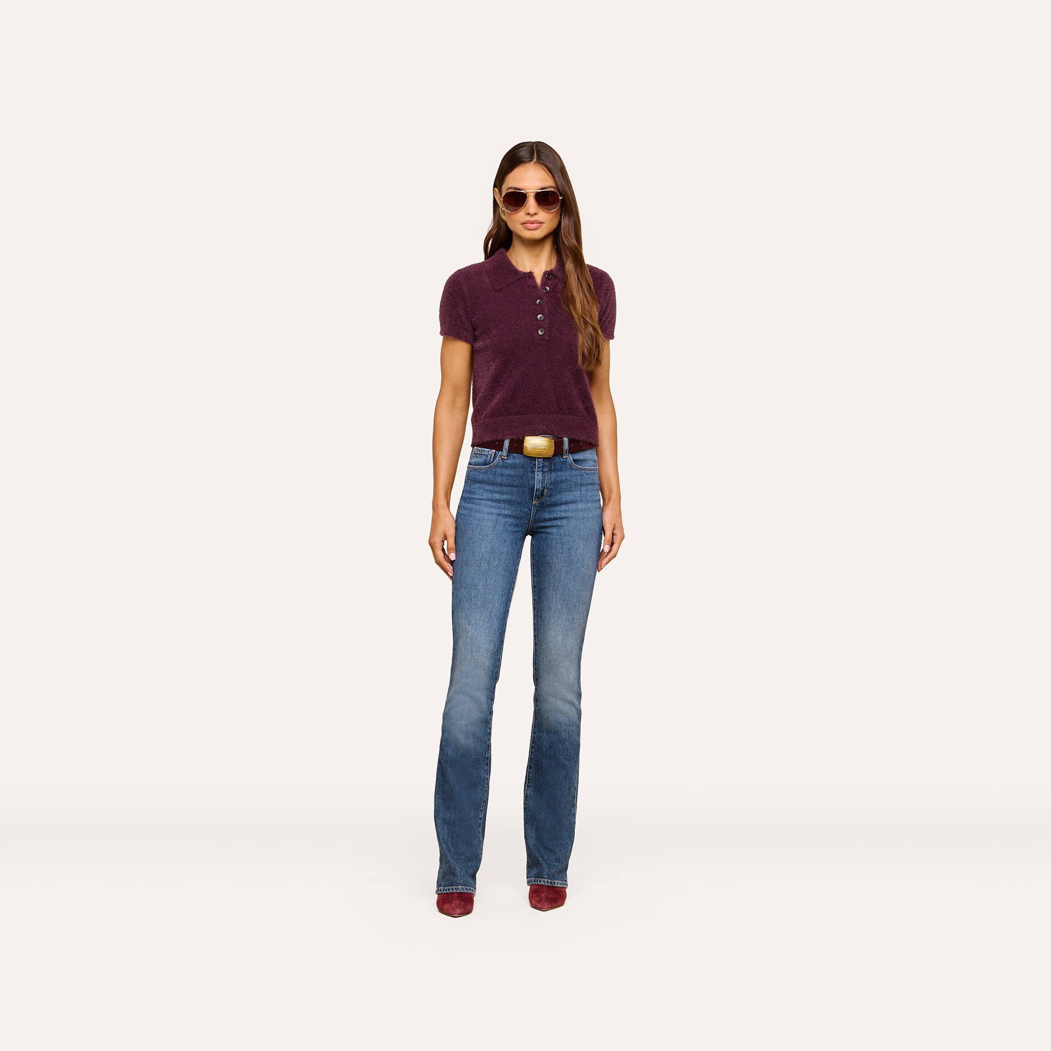 A woman stands facing forward against a plain white background, wearing sunglasses, blue jeans with a belt, red shoes, and the Cirrus Cropped Polo in maroon.