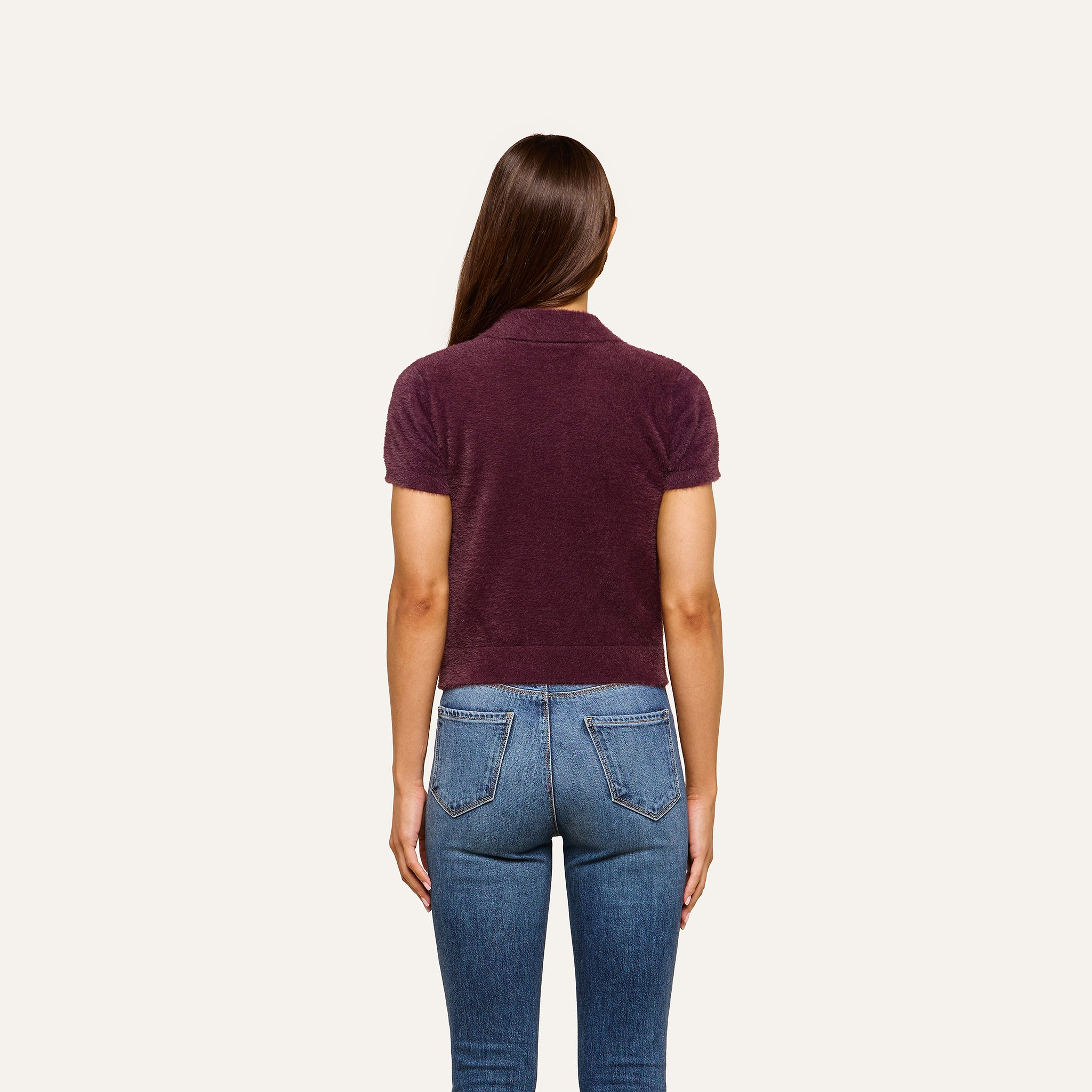 A person with long brown hair, wearing the Cirrus Cropped Polo in a soft knit along with blue jeans, stands facing away from the camera against a plain light background.