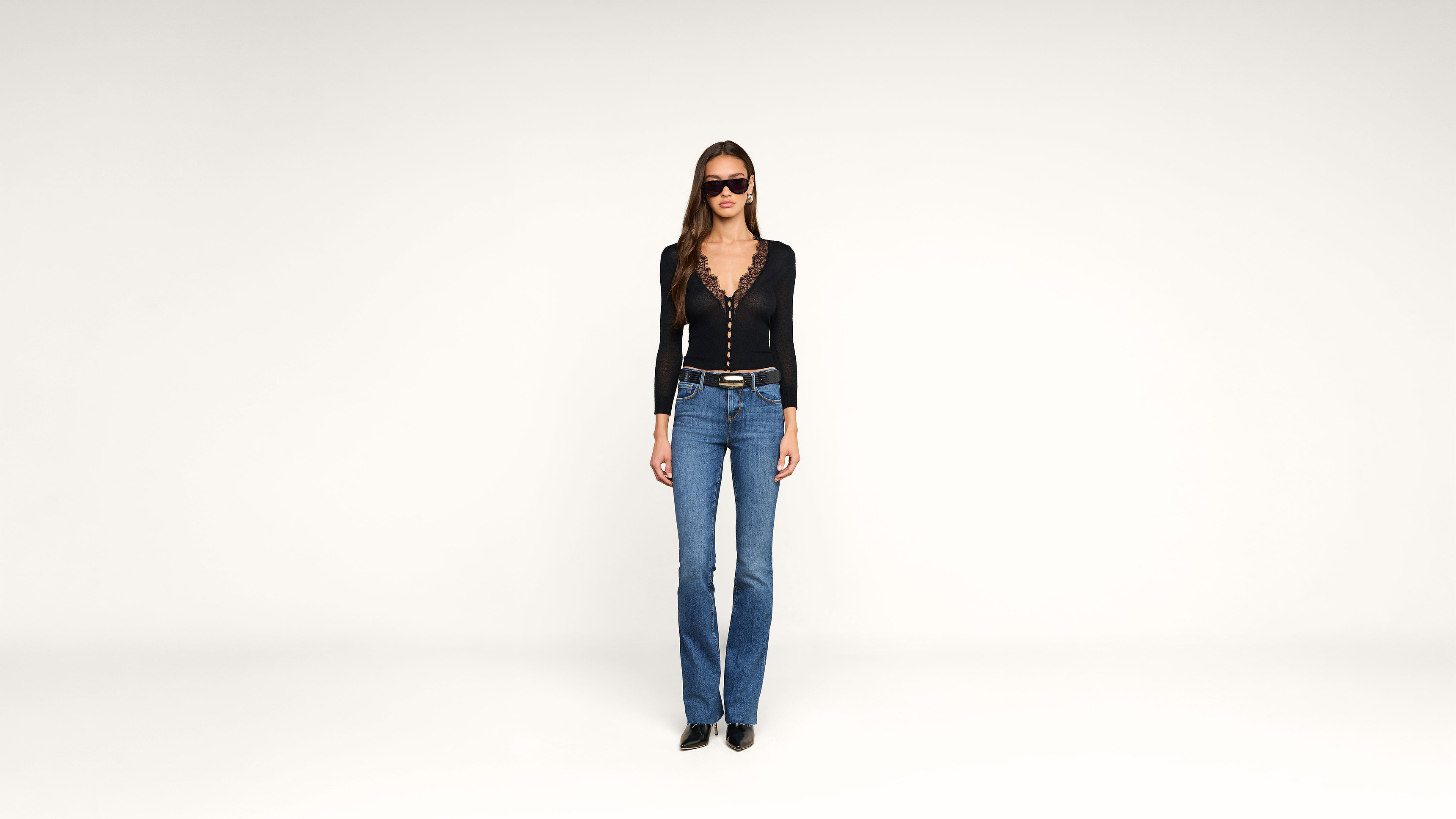 Woman stands against a plain white background, wearing sunglasses, a black long-sleeve top with a deep neckline, blue jeans, black shoes, and a black belt.