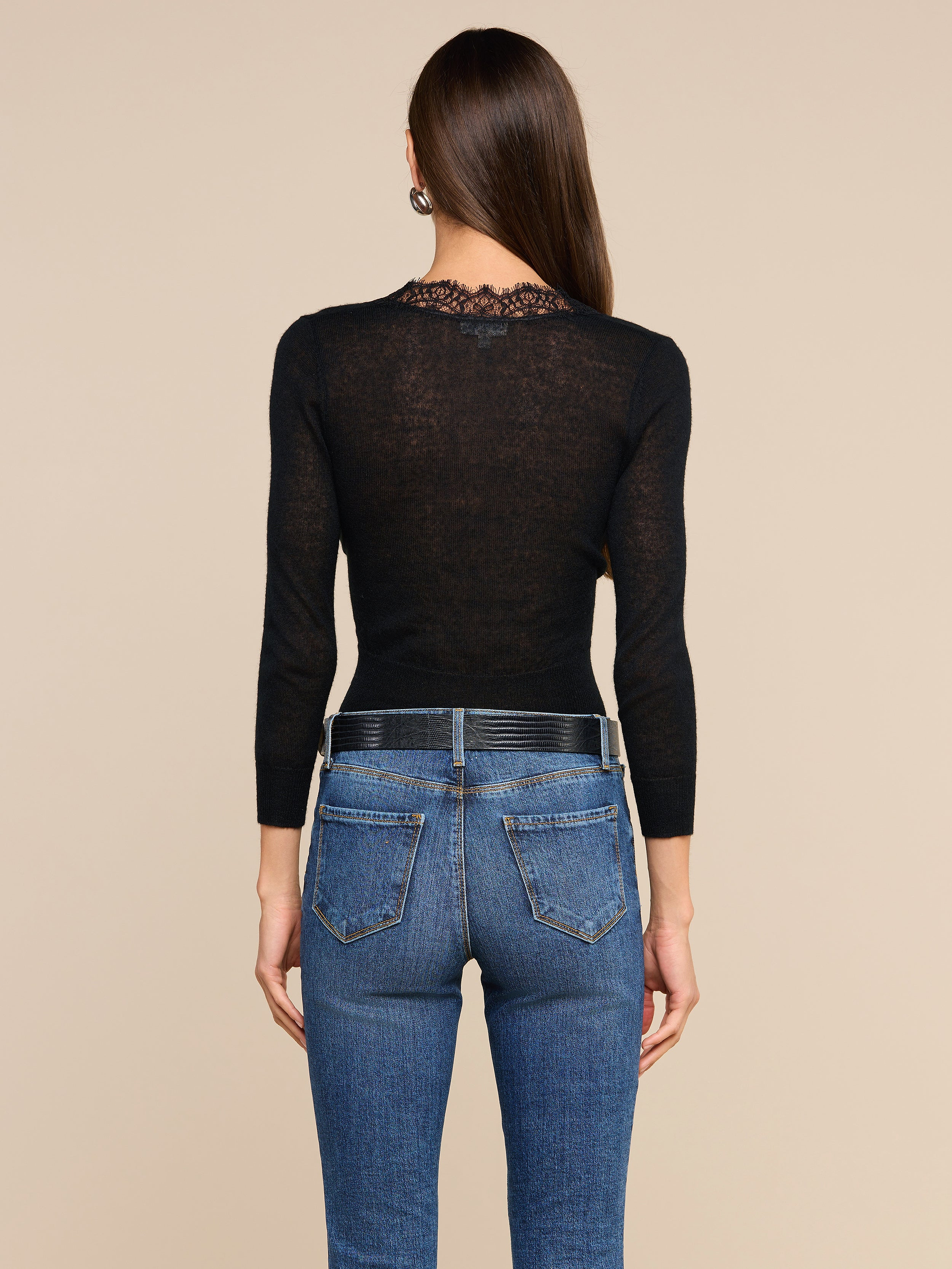 A person with long brown hair stands facing away, wearing the Eluna Cardigan Top—a black wool-blend long-sleeve top featuring a lace inset at the neckline—paired with blue jeans with visible back pockets, against a plain light background.