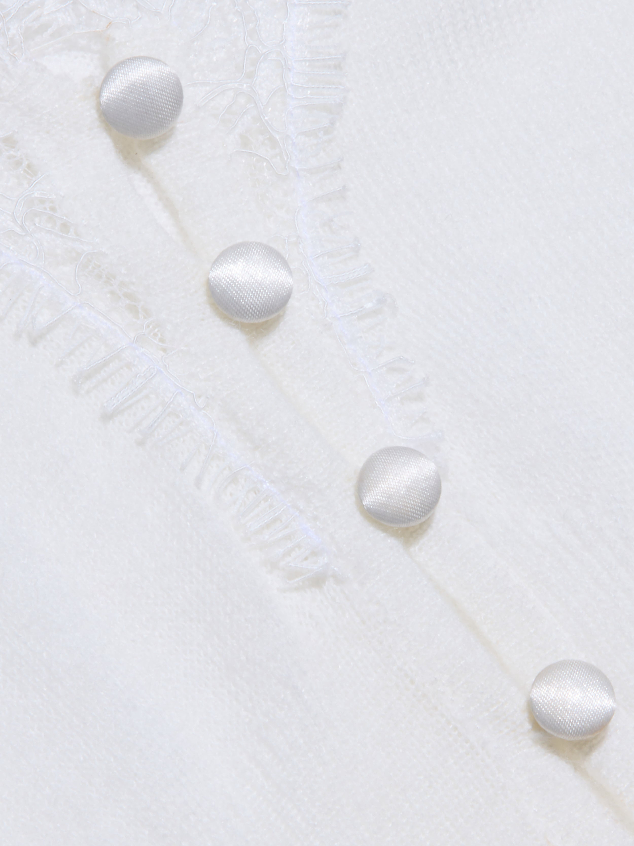 Primo piano del tessuto di maglia bianco e leggero come una piuma dellEluna Cardigan Top, con una texture sottile. Quattro bottoni rotondi ricoperti di tessuto sono disposti in diagonale e vicino ai bottoni sono presenti delicati dettagli in pizzo.