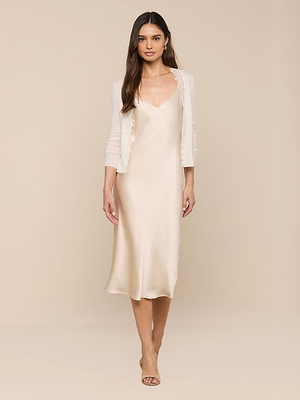 A woman with long brown hair faces forward in a cream satin slip dress and the Eluna Cardigan Top, paired with open-toe heels, standing against a beige background.