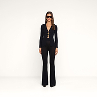 A woman stands against a plain white background wearing the fitted black Kaye Cardigan Top with gold clasps, paired with black flared pants, black shoes, and sunglasses. She looks straight ahead with her arms relaxed at her sides, creating a sleek travel set look.