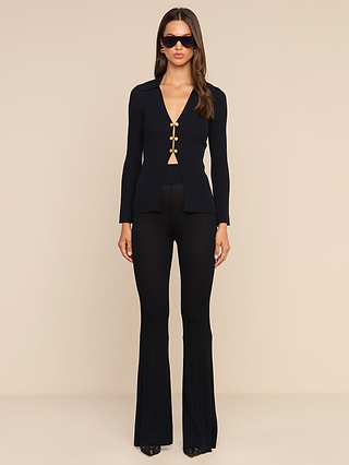 A woman stands facing forward against a beige background, wearing the Kaye Cardigan Top—a black ribbed knit buttoned top—paired with flared black pants, sunglasses, and loose hair for an effortlessly stylish travel set.
